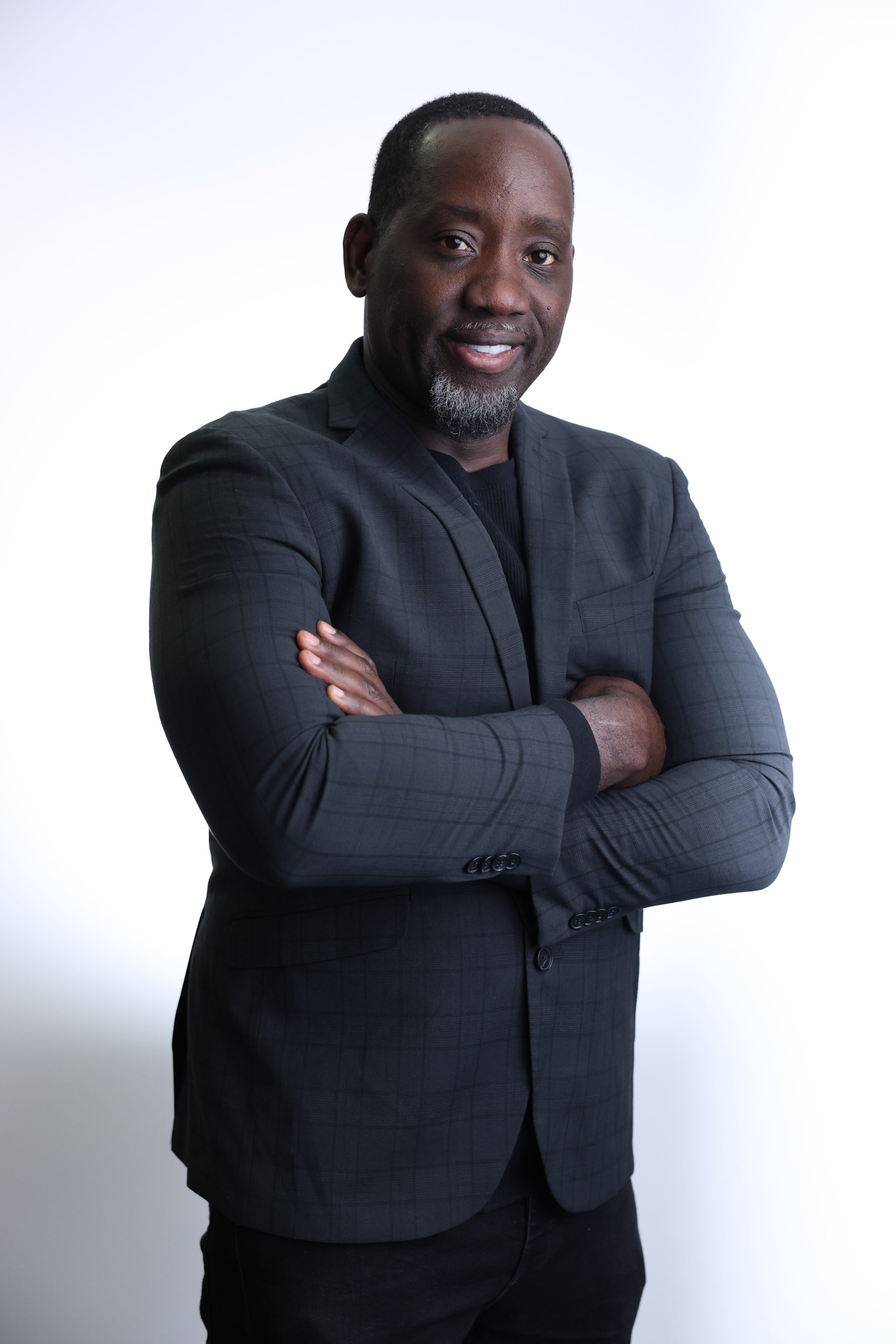 Smiling man in a dark blazer with arms crossed against a black background. He appears confident and approachable.