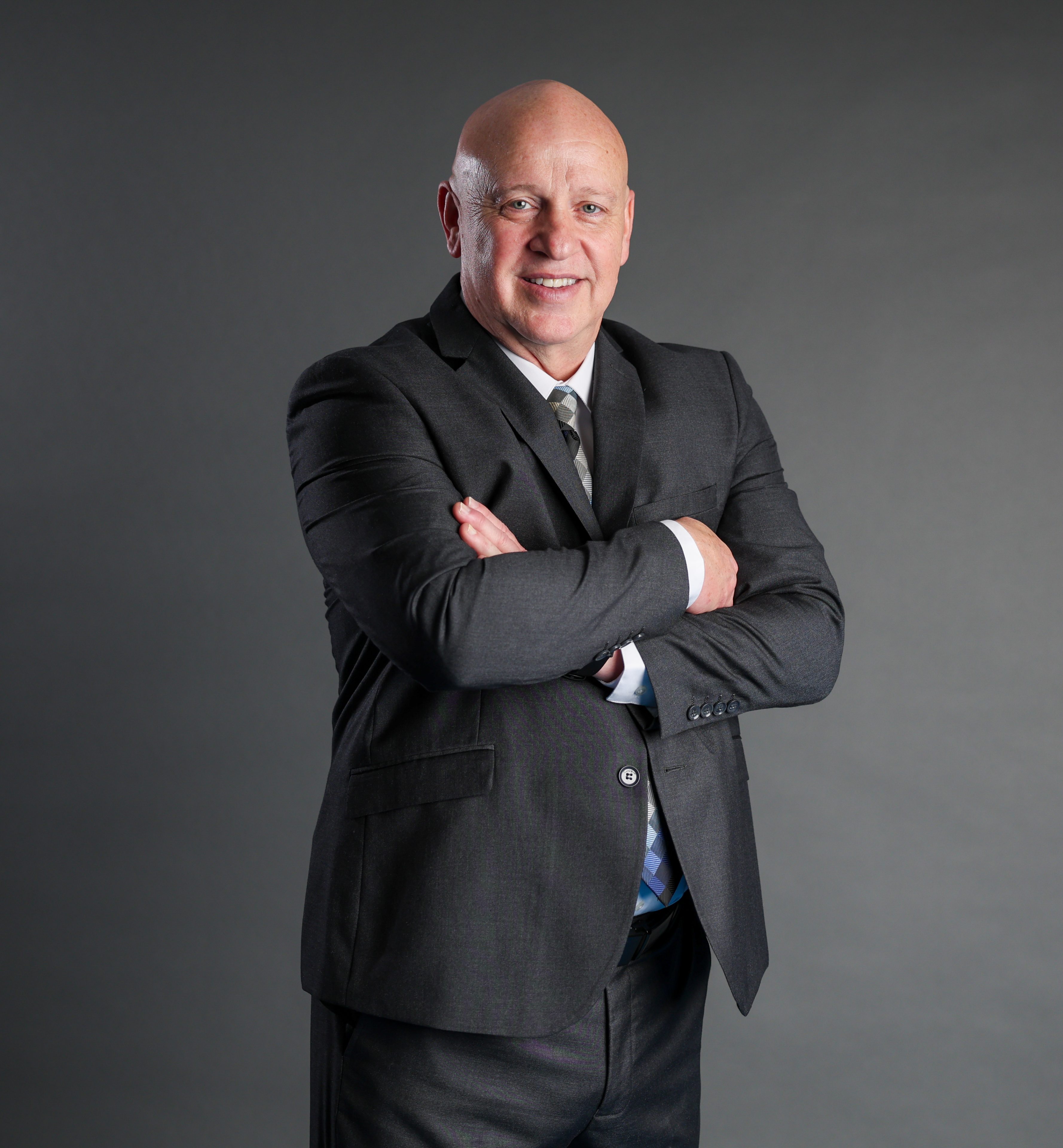 A confident bald man in a dark suit stands with arms crossed against a gray background, smiling warmly, conveying professionalism and approachability.