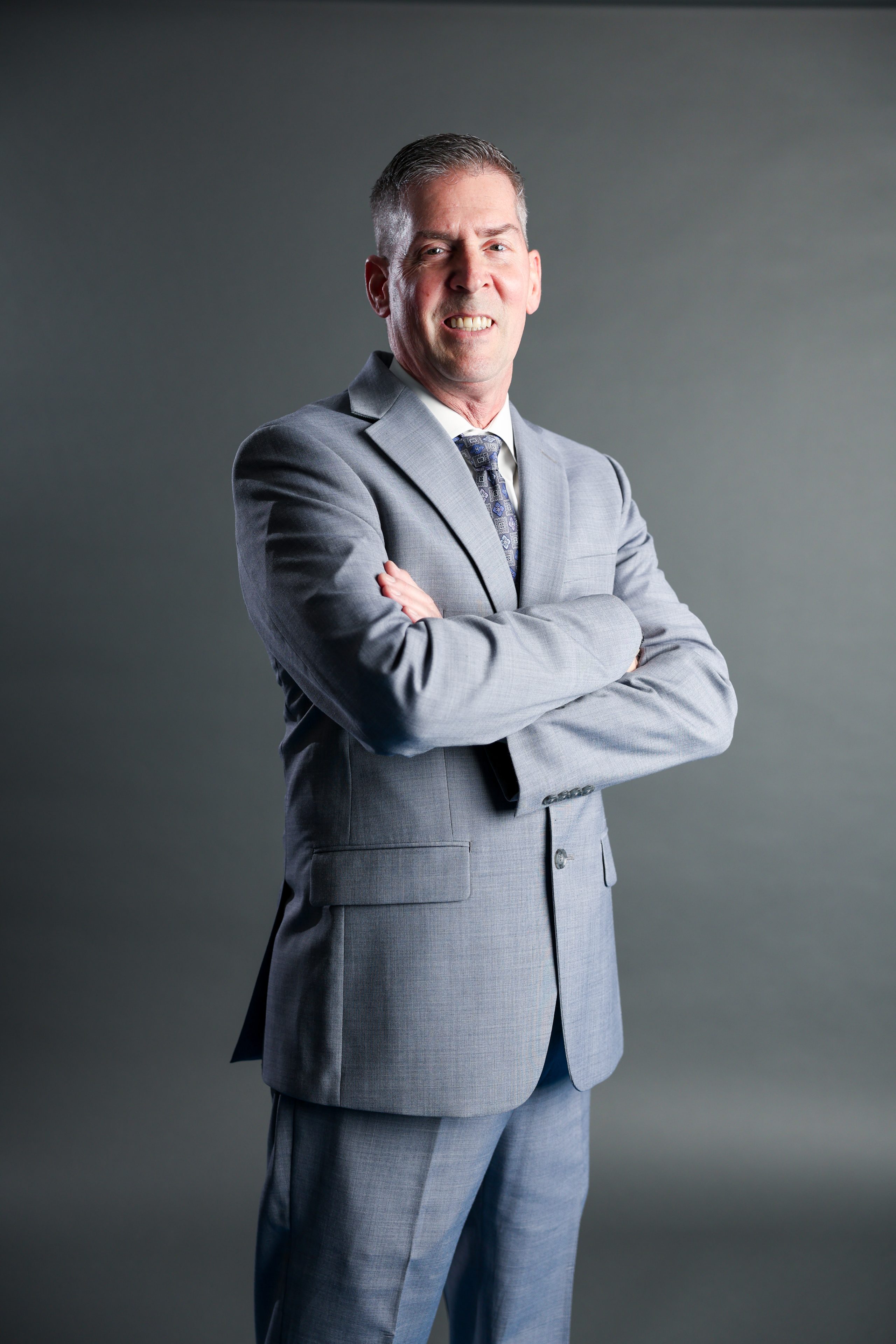 Man in a light gray suit stands confidently with arms crossed, smiling slightly. The background is plain gray, conveying a professional tone.