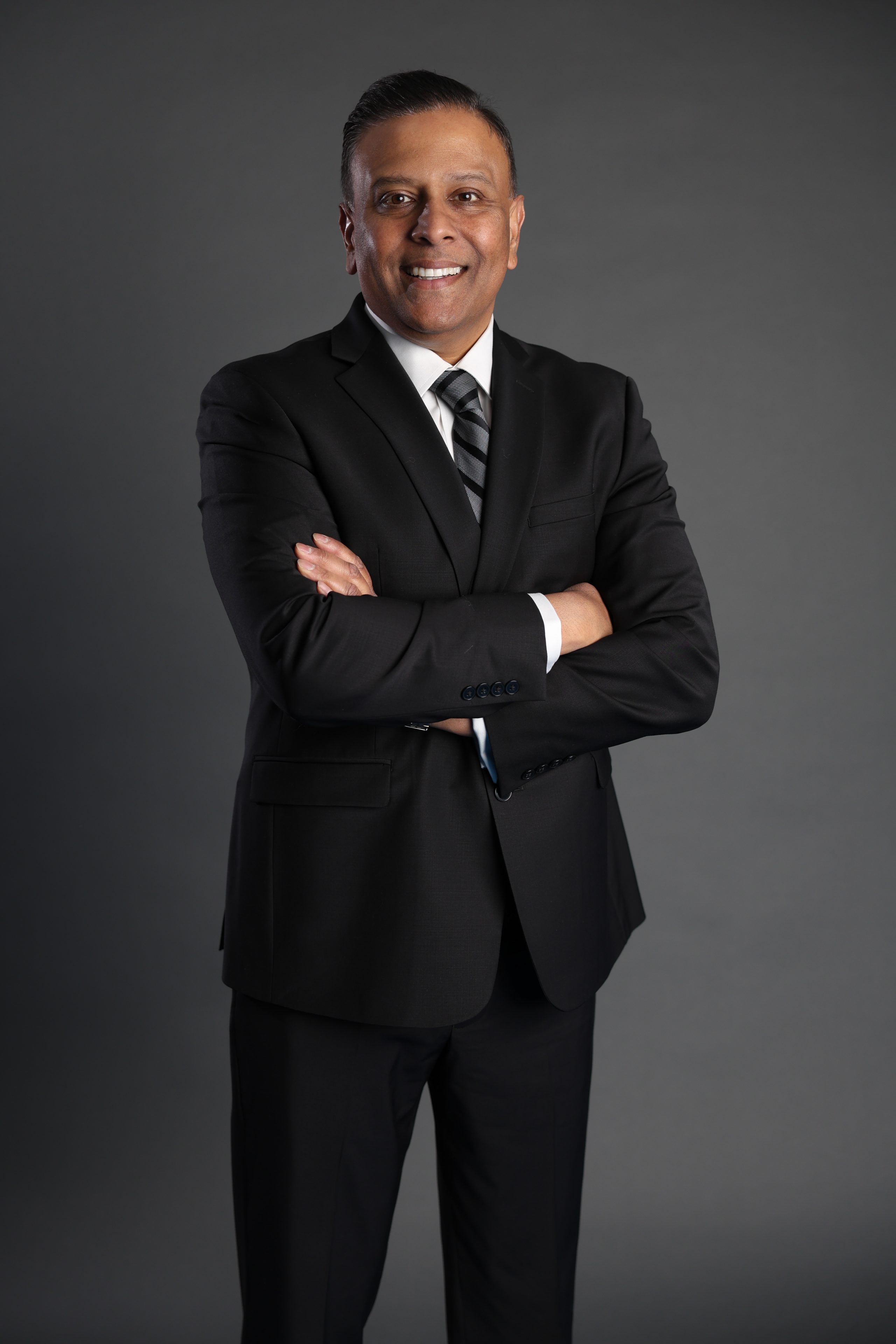 A confident man stands with arms crossed, wearing a black suit and tie, against a plain gray background, conveying professionalism and poise.