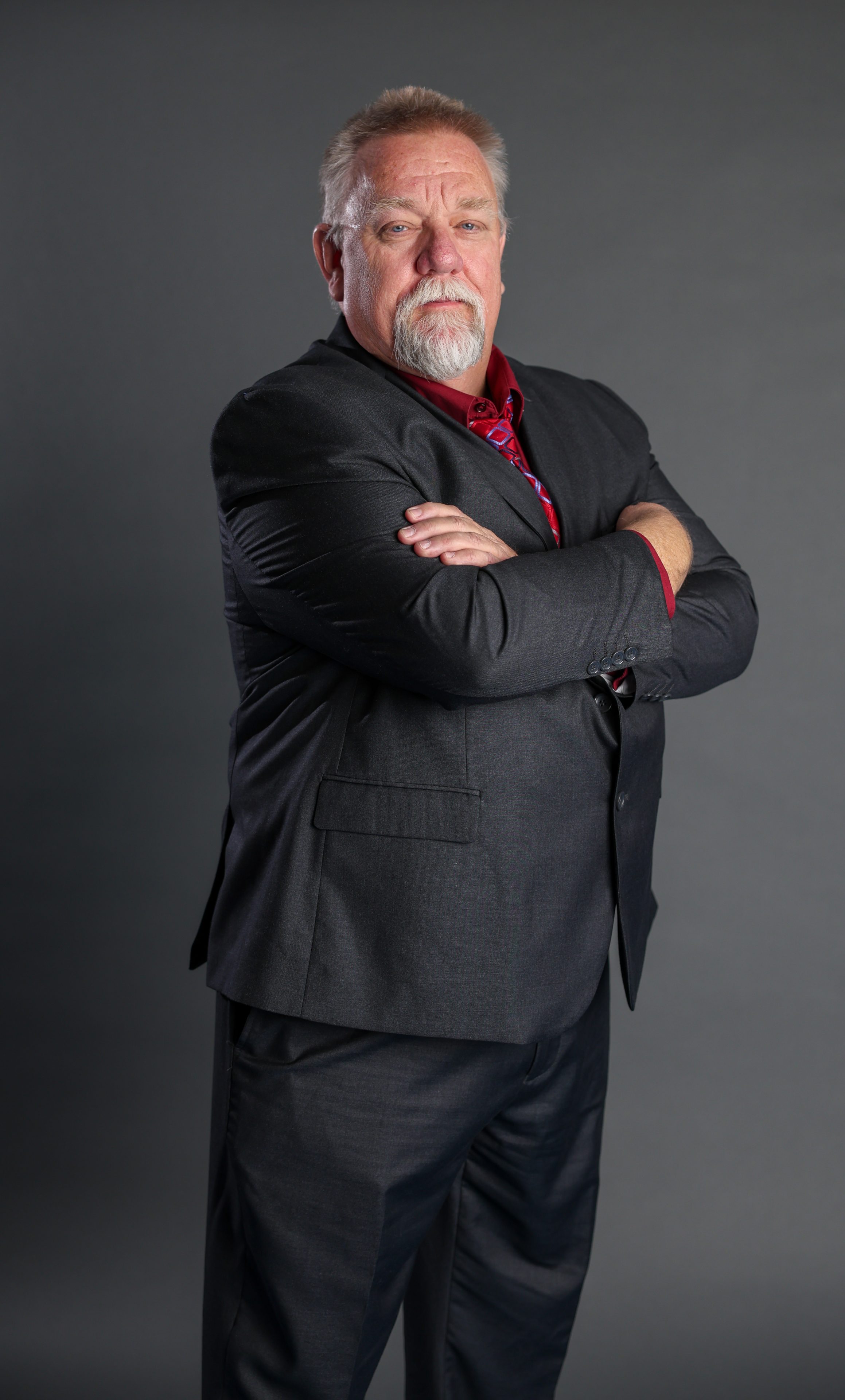 A stern-looking man with a gray beard stands confidently with arms crossed, wearing a black suit and red shirt. Gray background, professional tone.
