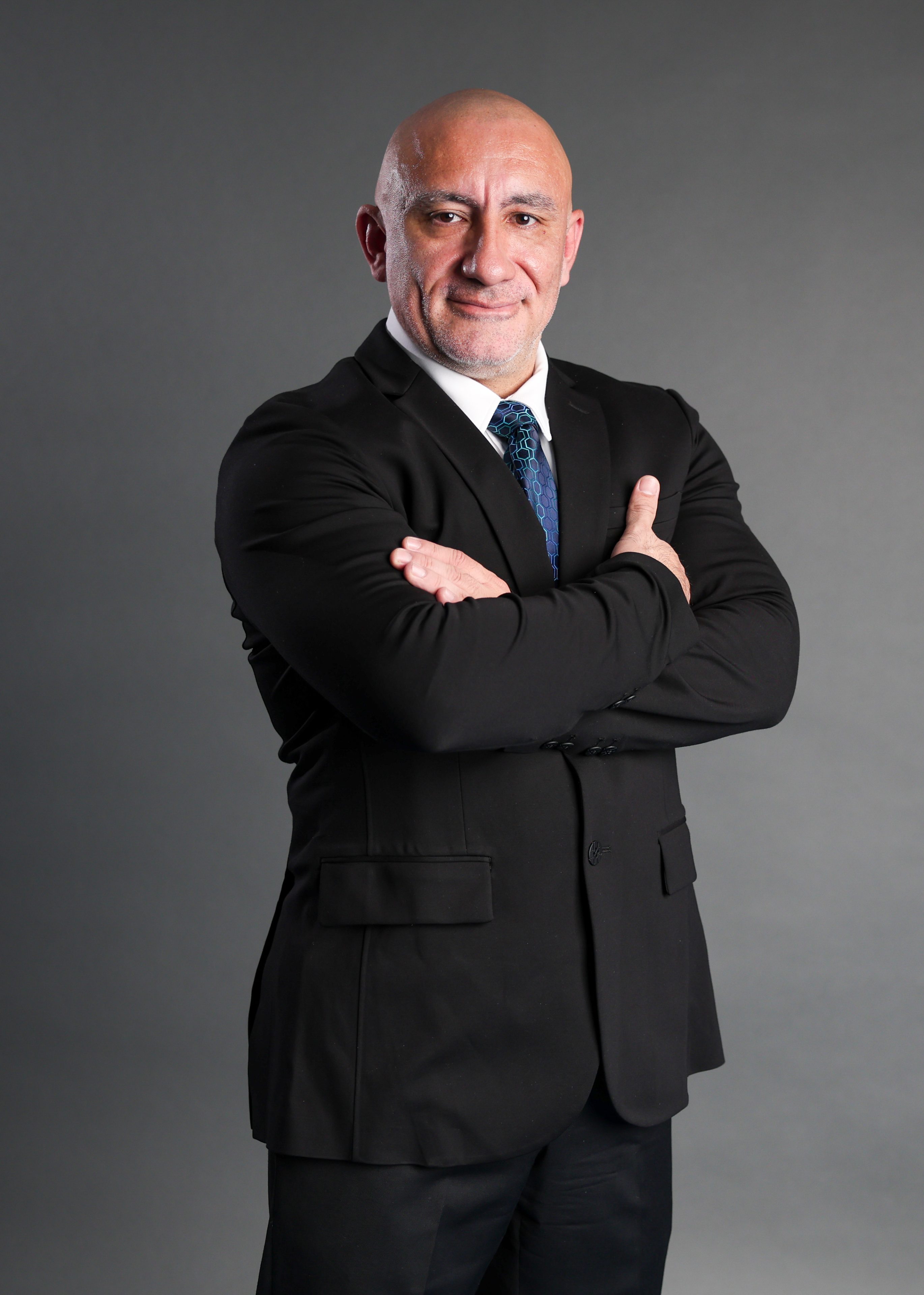 A bald man in a black suit and blue tie stands confidently with arms crossed against a gray background, conveying professionalism and authority.