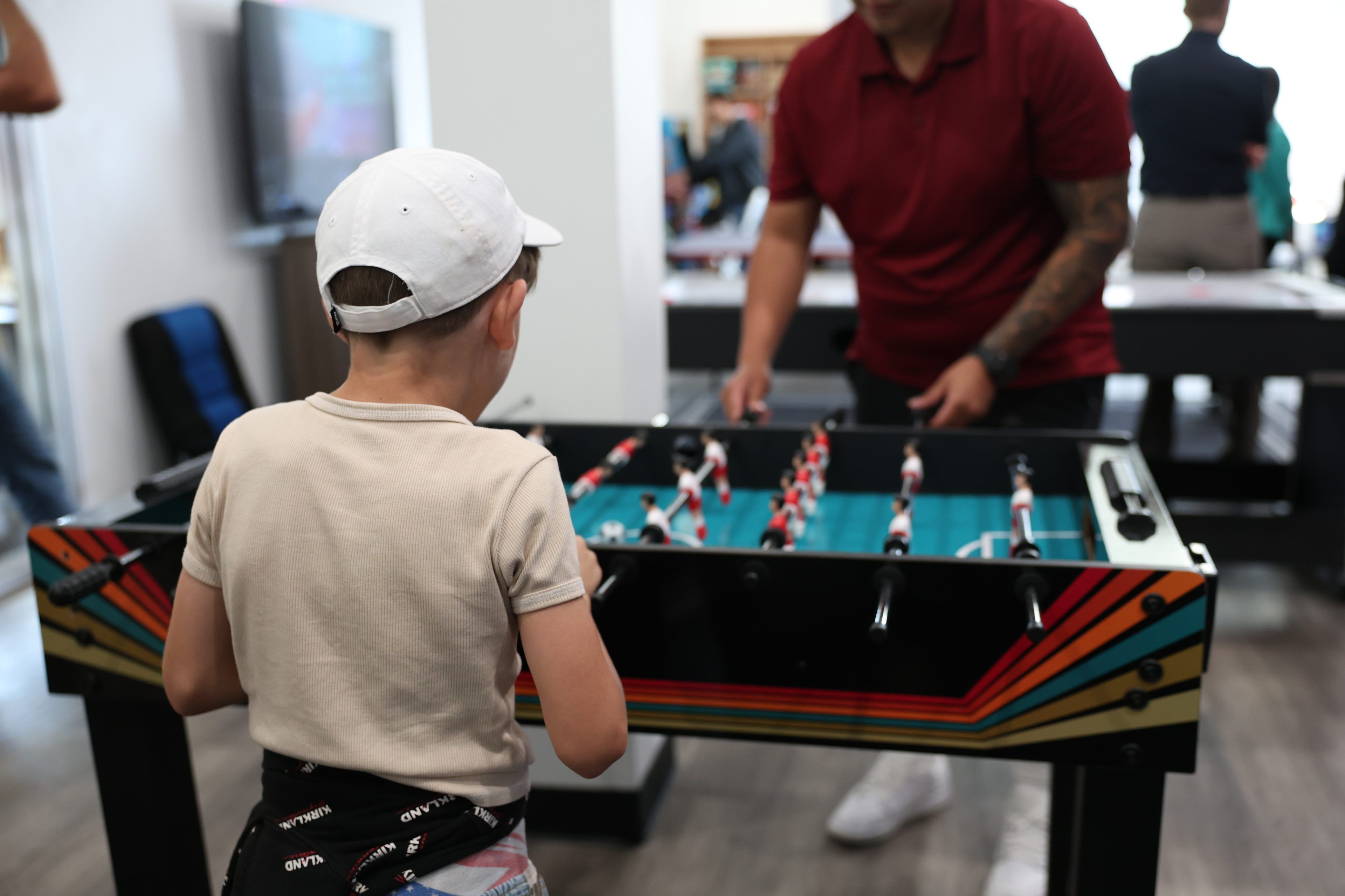 A young boy and an adult are playing foosball in a lively indoor setting. The boy, wearing a white cap, is focused, while the room has a relaxed, social atmosphere.