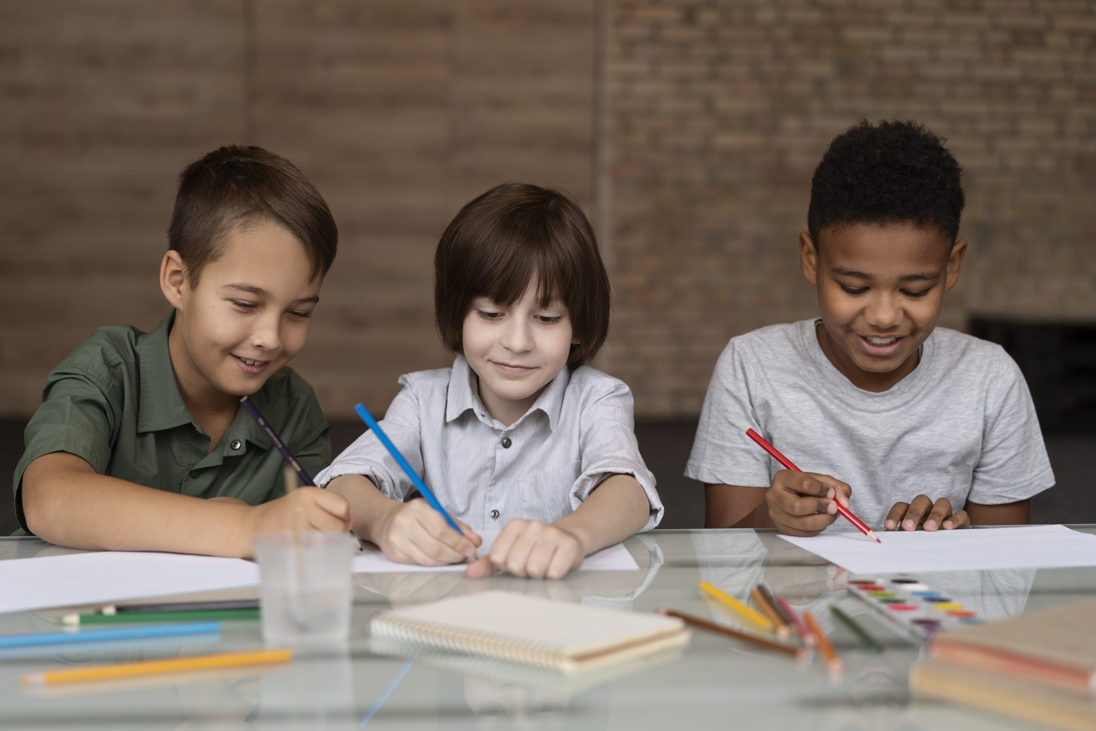 Three children, smiling and focused, are drawing with colored pencils at a table. Their art supplies and a brick wall are visible in the background.