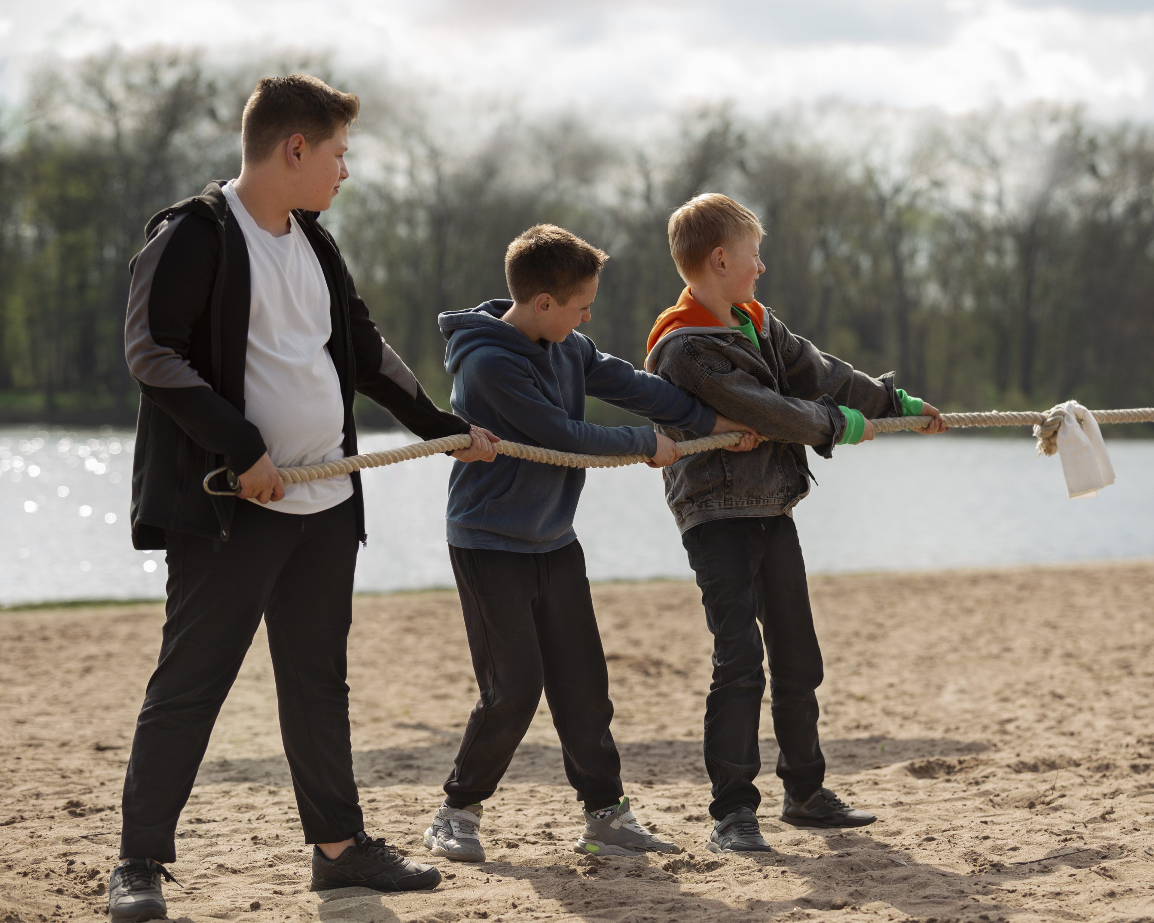 Three boys play tug-of-war on a sandy beach. They pull a rope, appearing determined and focused, with a lake and trees in the background.