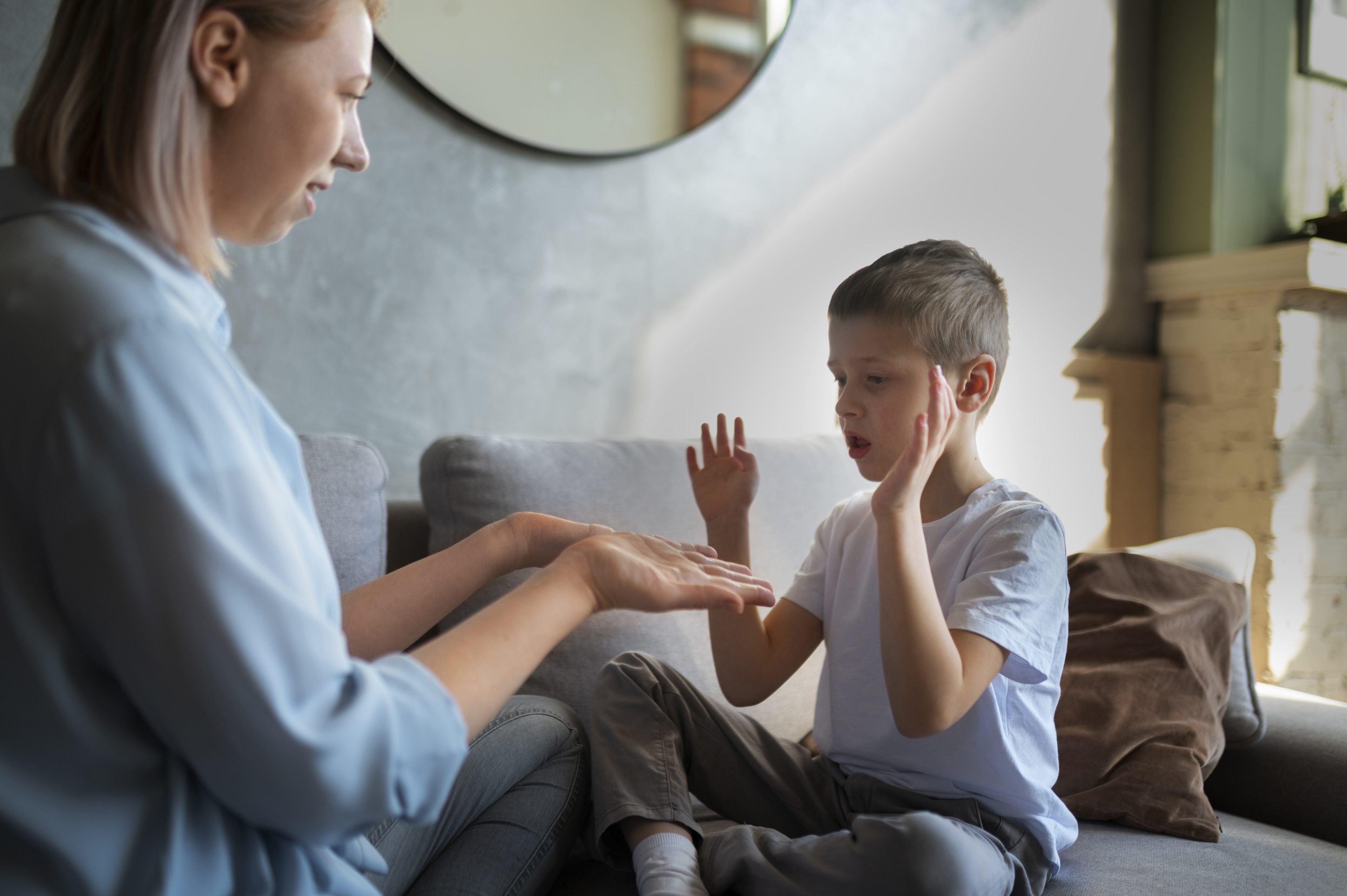 A woman and a boy sit on a couch, engaging in interactive communication. The boy raises his hands expressively, conveying focus and connection.