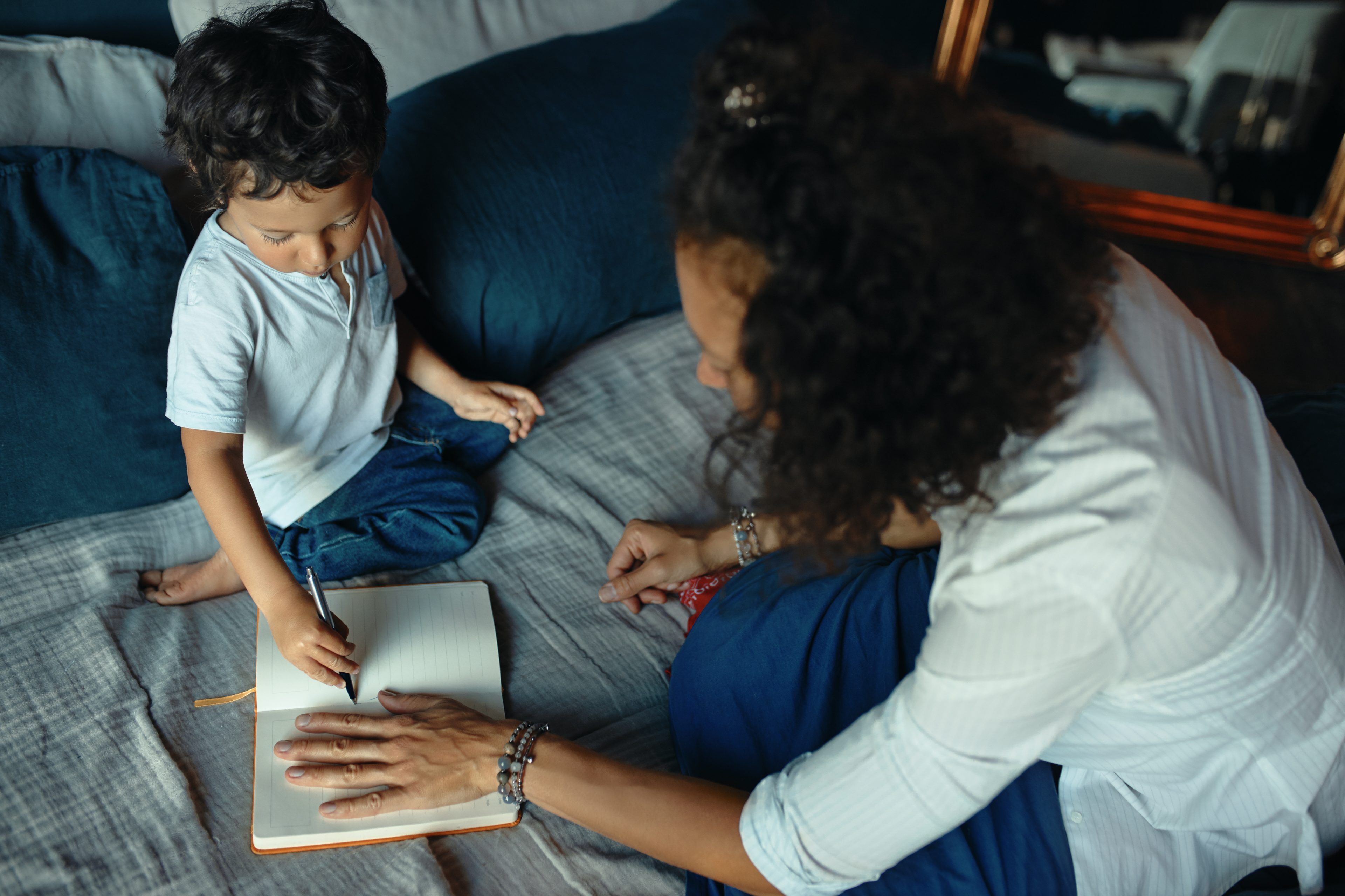 High angle view of young dark skinned woman with curly hair sitting on bed with hand on sheet in copybook, little boy drawing, tracing outline of her palm. Mom spending day with son at home