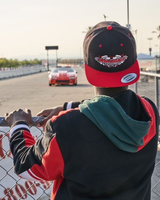 A person in a red and black jacket and cap watches a red race car zoom on a sunny track. The scene conveys excitement and anticipation at a car event.