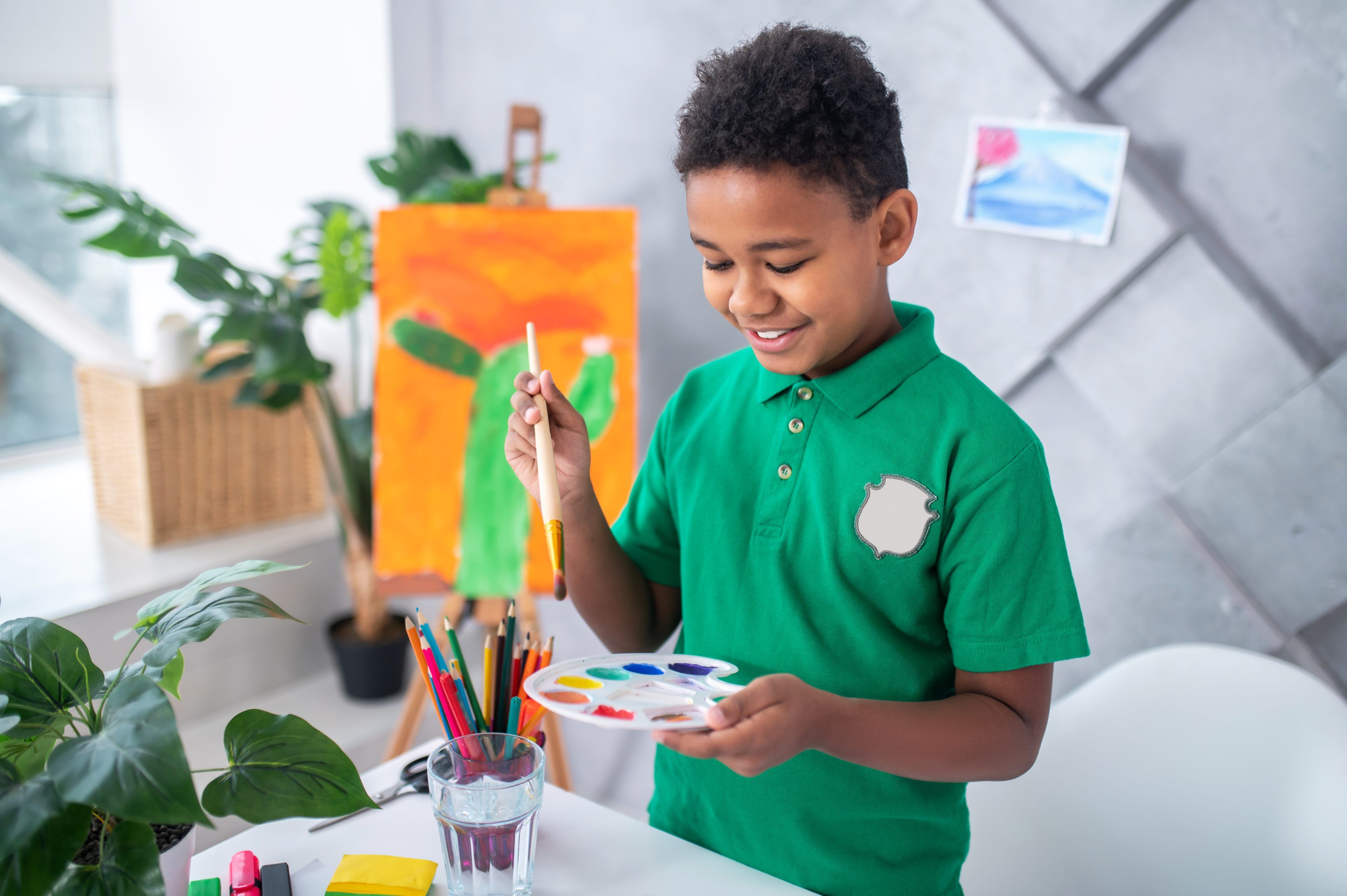 A smiling child in a green shirt holds a paintbrush and palette. Behind them is a colorful painting on an easel. The room feels bright and creative.