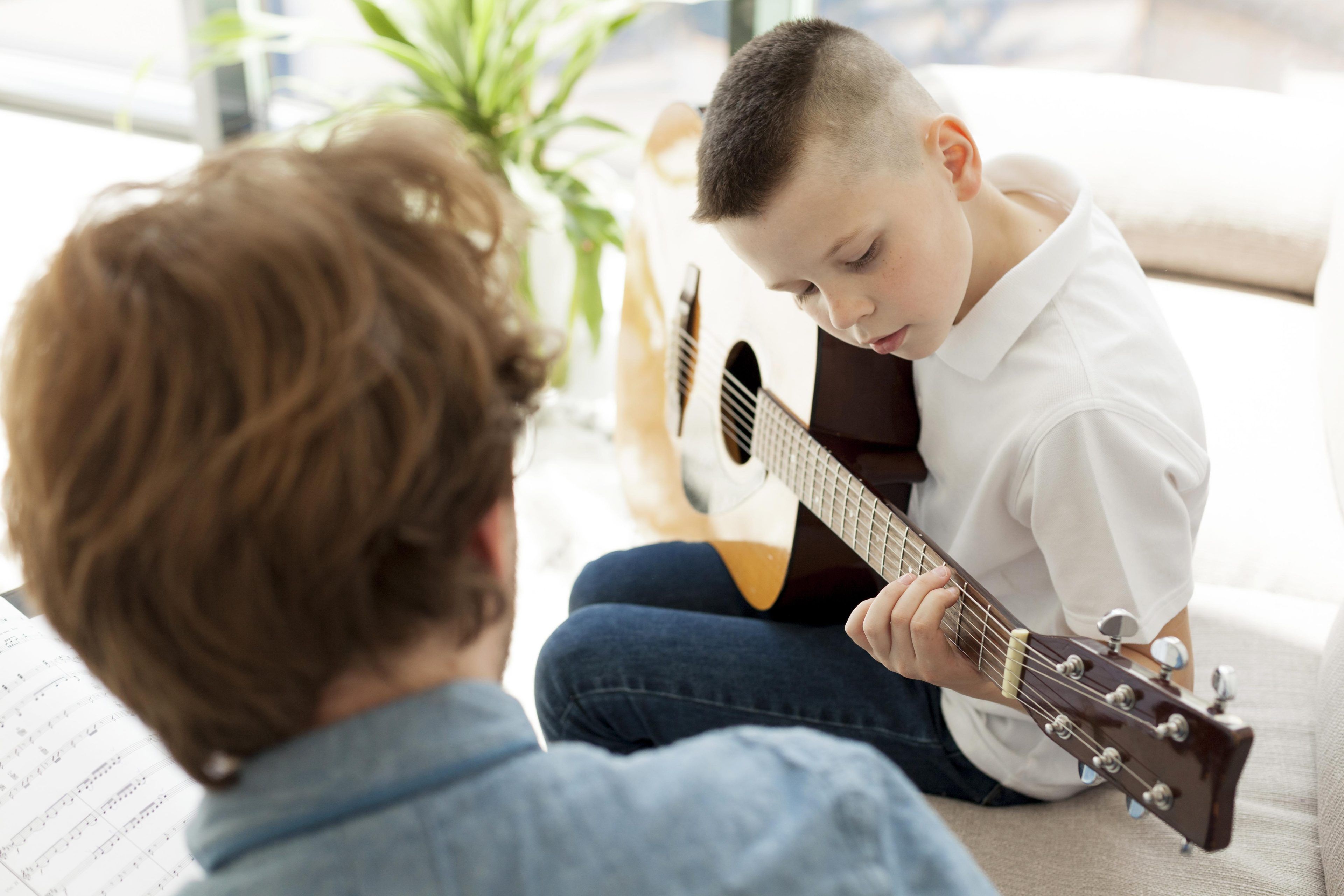 A young boy in a white shirt plays acoustic guitar, focusing intently. An adult with reddish hair guides him, sheet music visible. Bright, calm atmosphere.