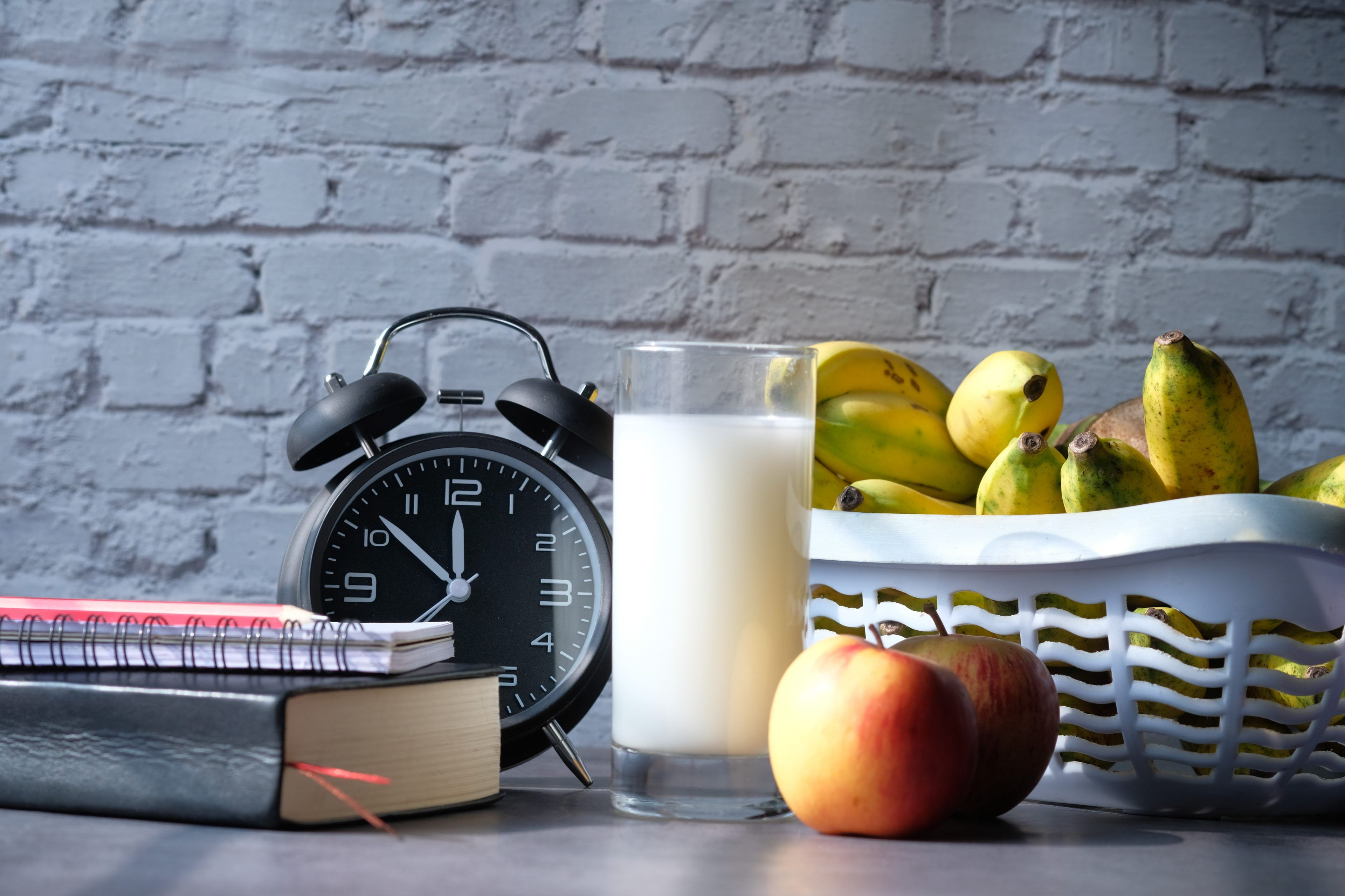 A black alarm clock, notebooks, a glass of milk, apples, and a basket of bananas are set against a gray brick wall, conveying a morning routine theme.