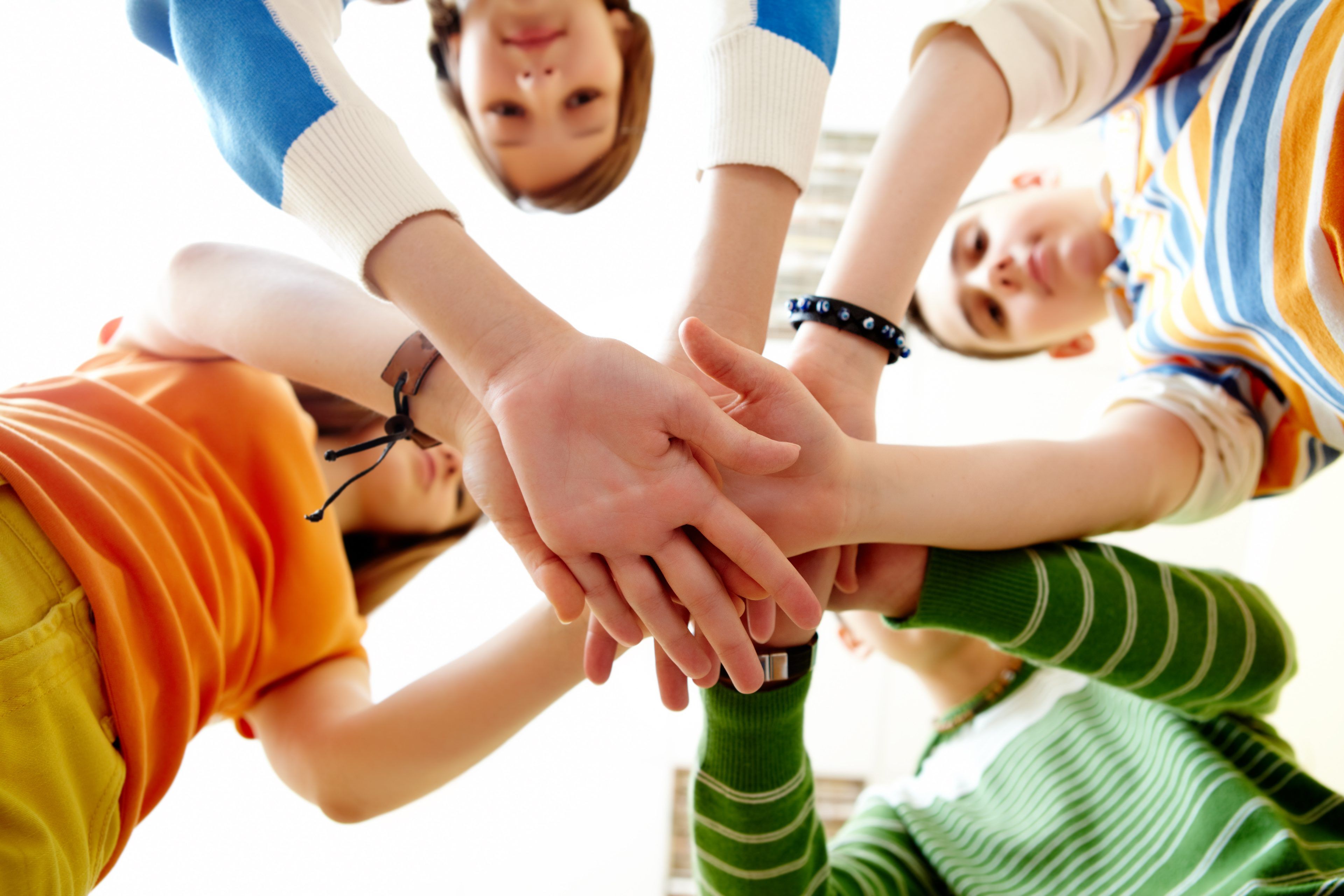 Four children standing in a circle, seen from below, place their hands together in the center, symbolizing teamwork and unity. They are smiling and wearing colorful clothing.