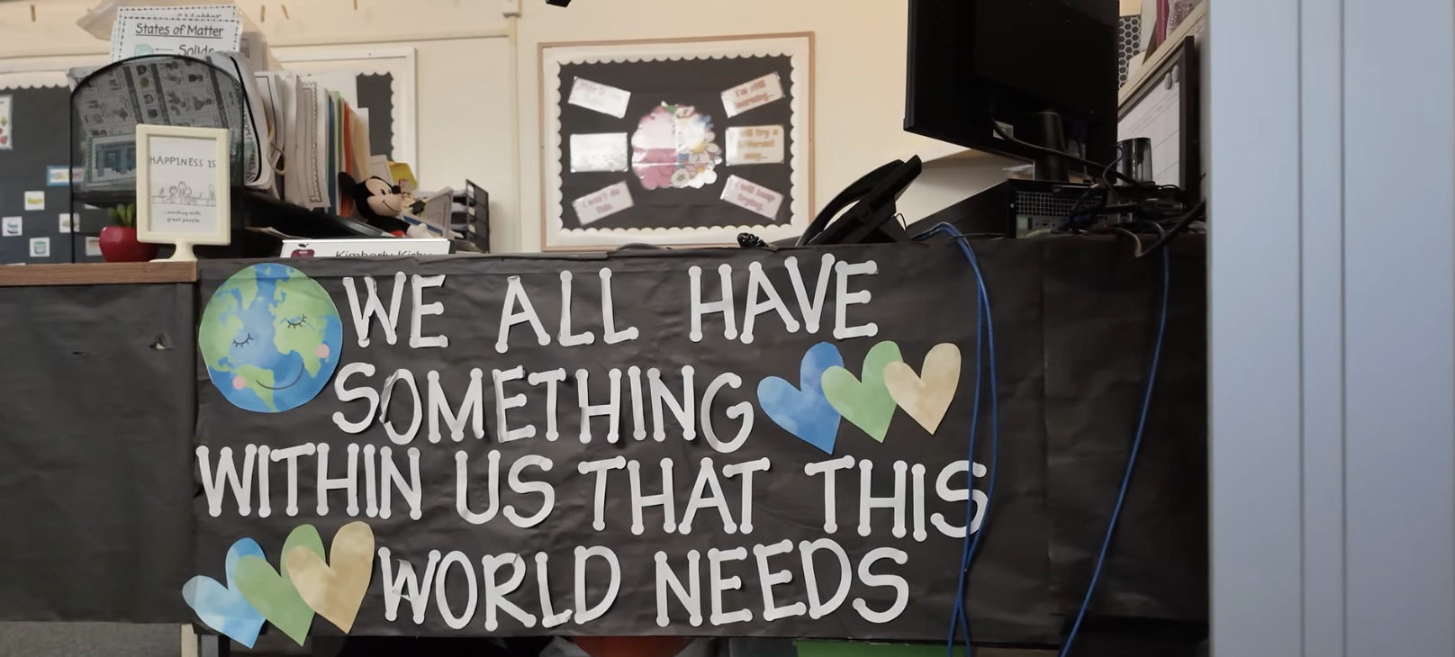 A classroom with a sign reading "We all have something within us that this world needs" on a wall. The sign features a globe and heart shapes, conveying a positive message.