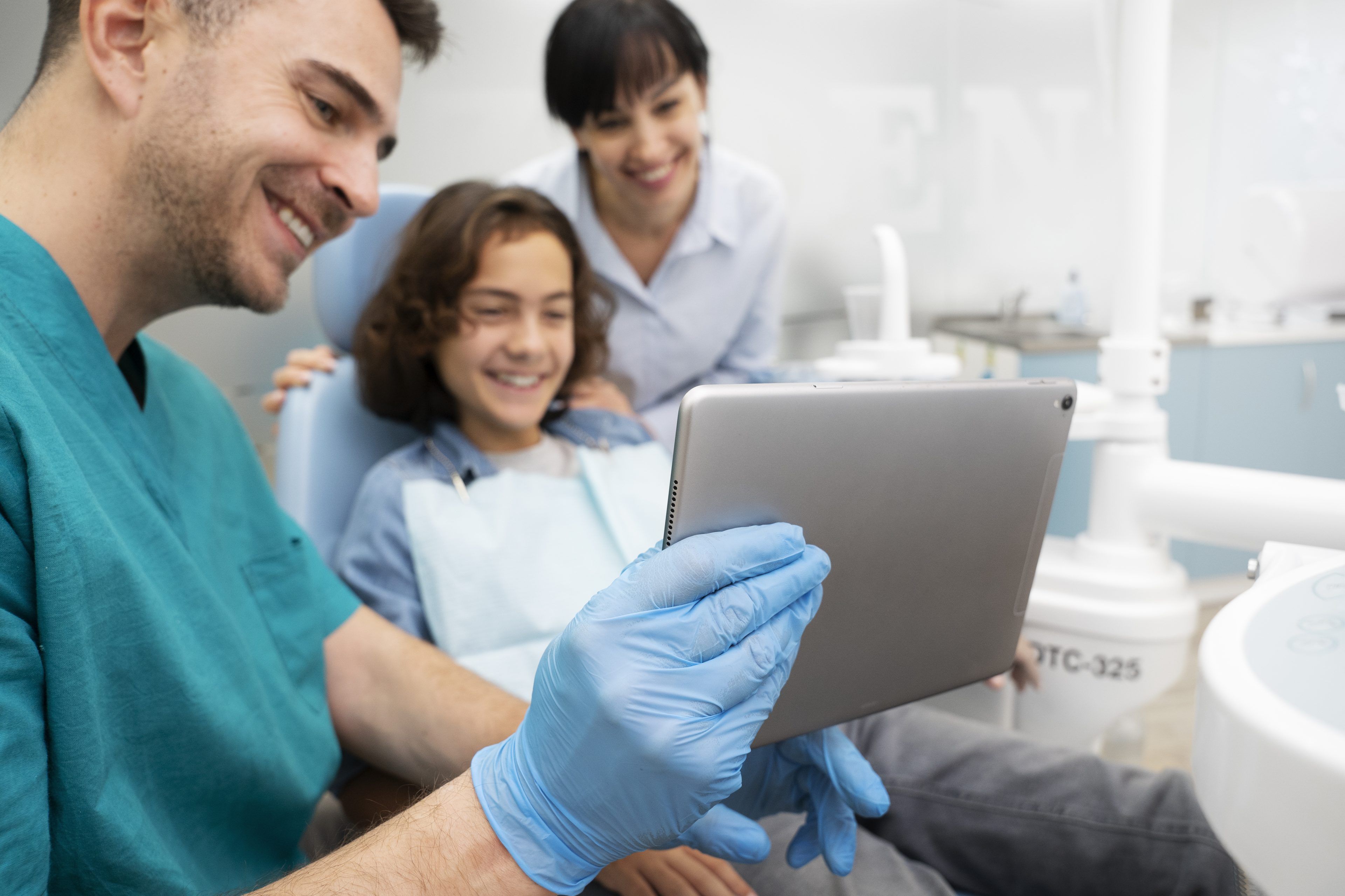 Smiling dentist shows a young patient and their parent something on a tablet. The child wears a dental apron, creating a friendly, reassuring atmosphere.