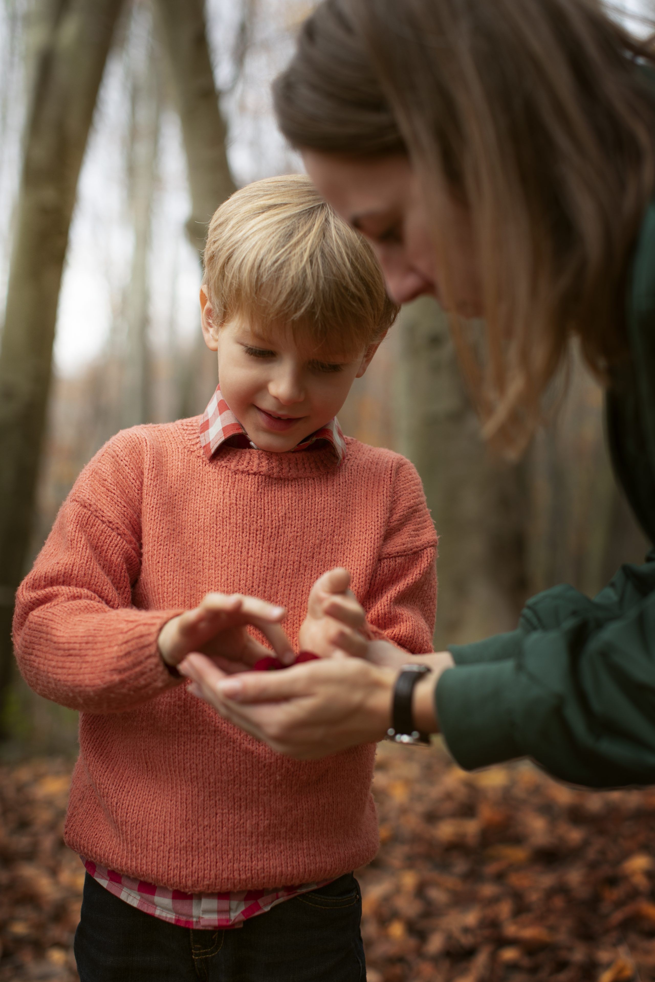 A young boy in an orange sweater examines something in his hand, assisted by an adult in a green jacket. They are in a forest with autumn leaves, conveying curiosity.