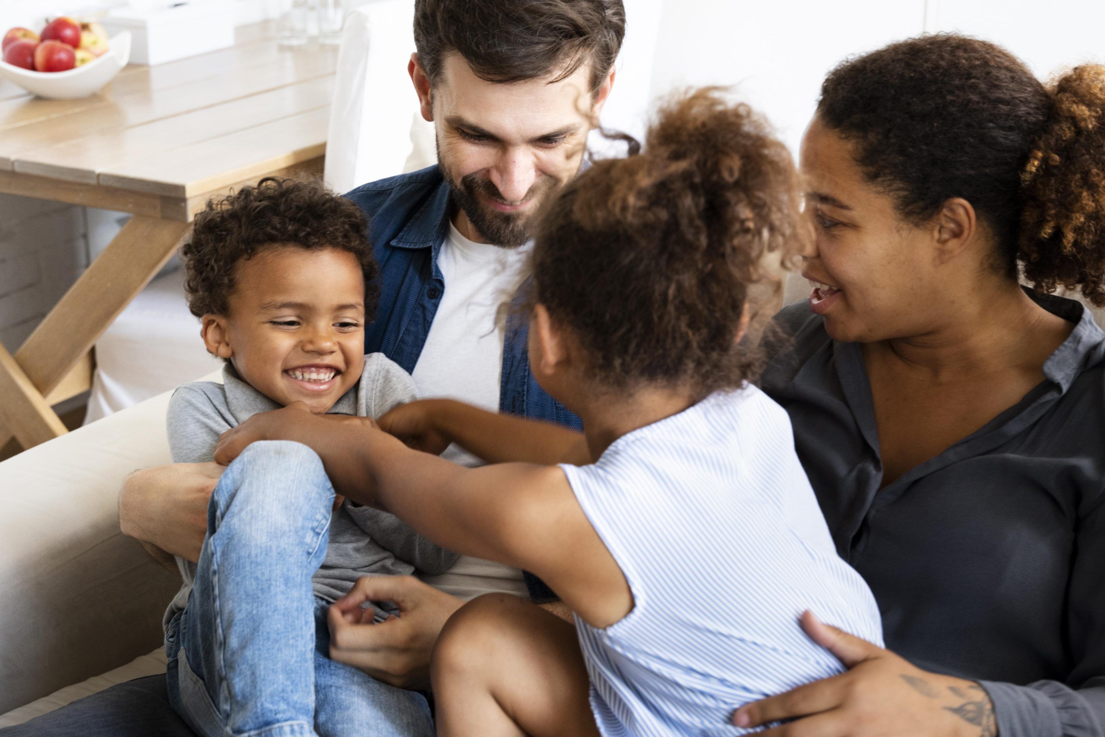 A happy family of four sits on a couch. The parents watch with smiles as the kids, a boy and a girl, laugh and playfully interact. The setting is warm and homey.