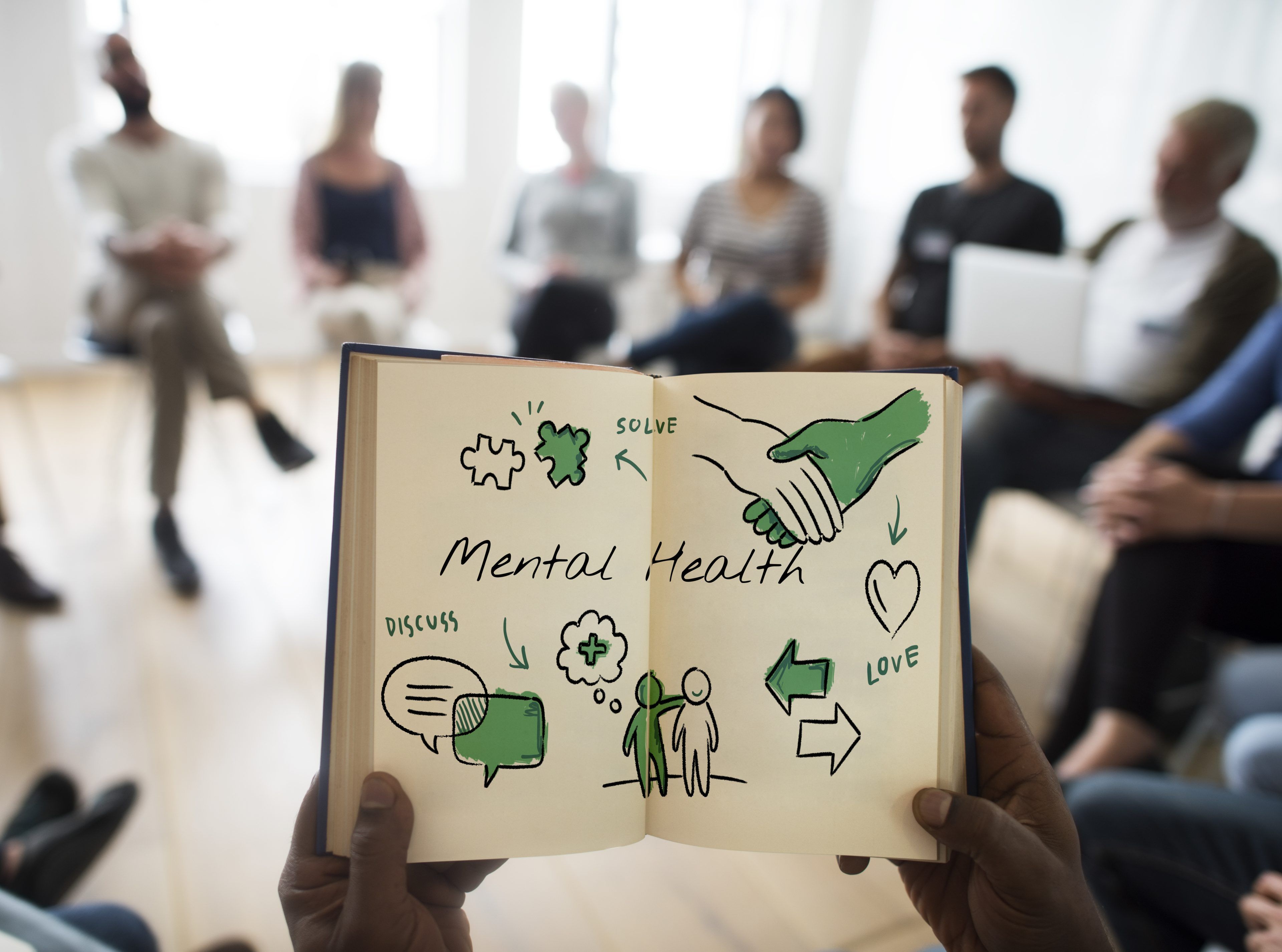 A book titled "Mental Health" with drawings of puzzle pieces, speech bubbles, and hands shakes is held up. People sit in a circle in the background, suggesting a supportive group setting.