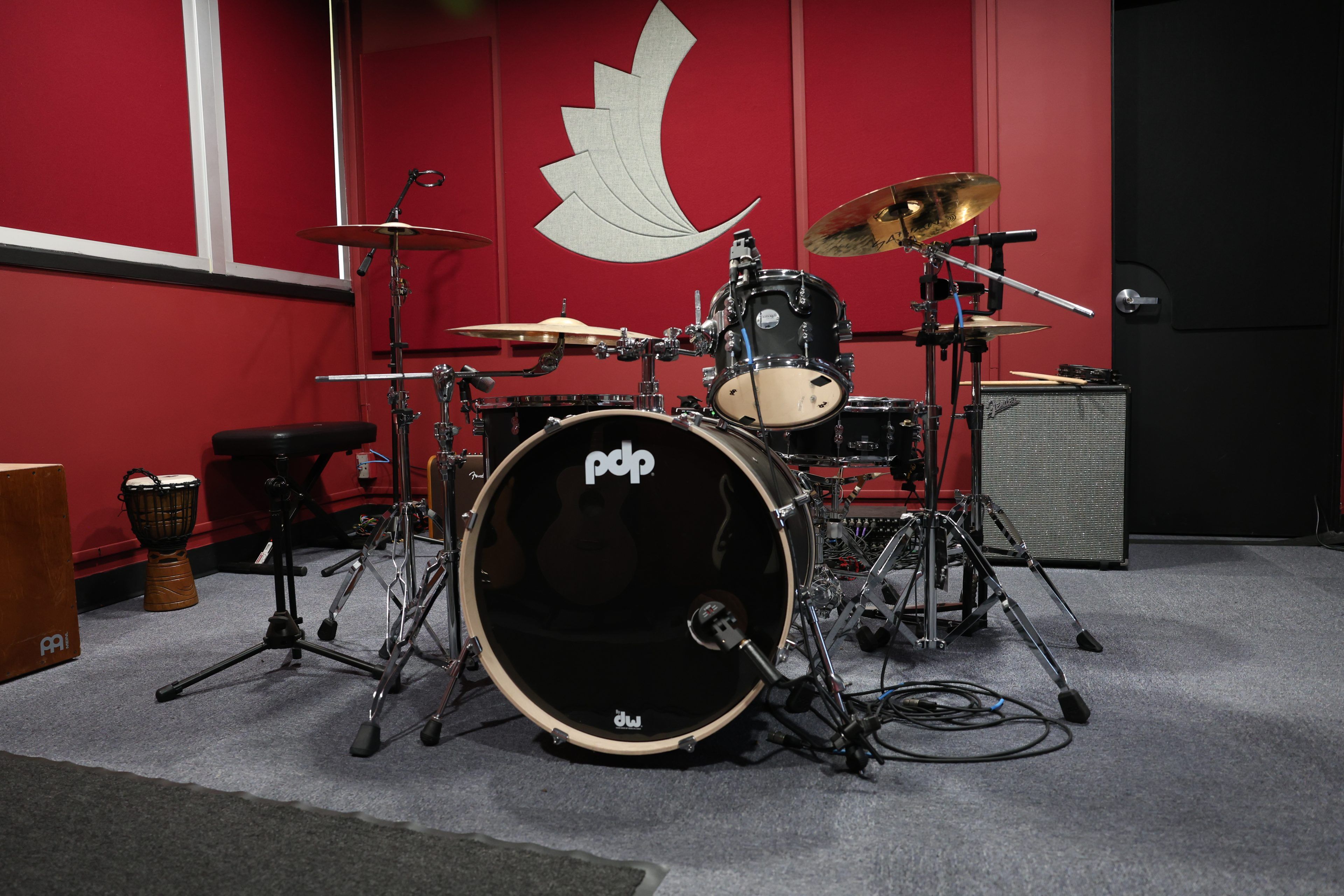 A drum set with a black PDP bass drum sits in a music studio with red walls and a white abstract design, evoking a professional, creative vibe.