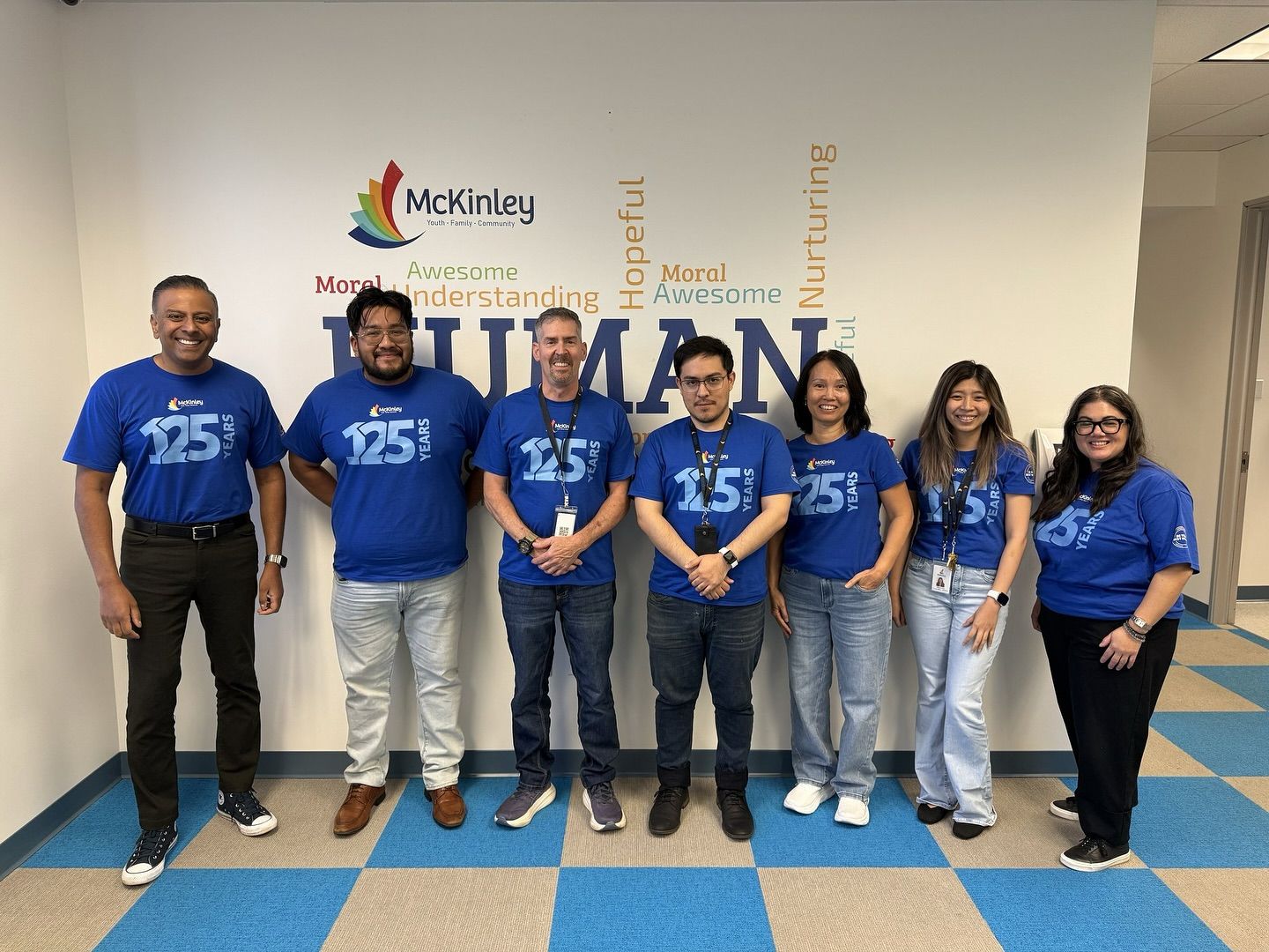 A group of eight people, smiling and wearing matching blue "25" shirts, stands in front of a wall with inspirational words. The mood is cheerful and celebratory.