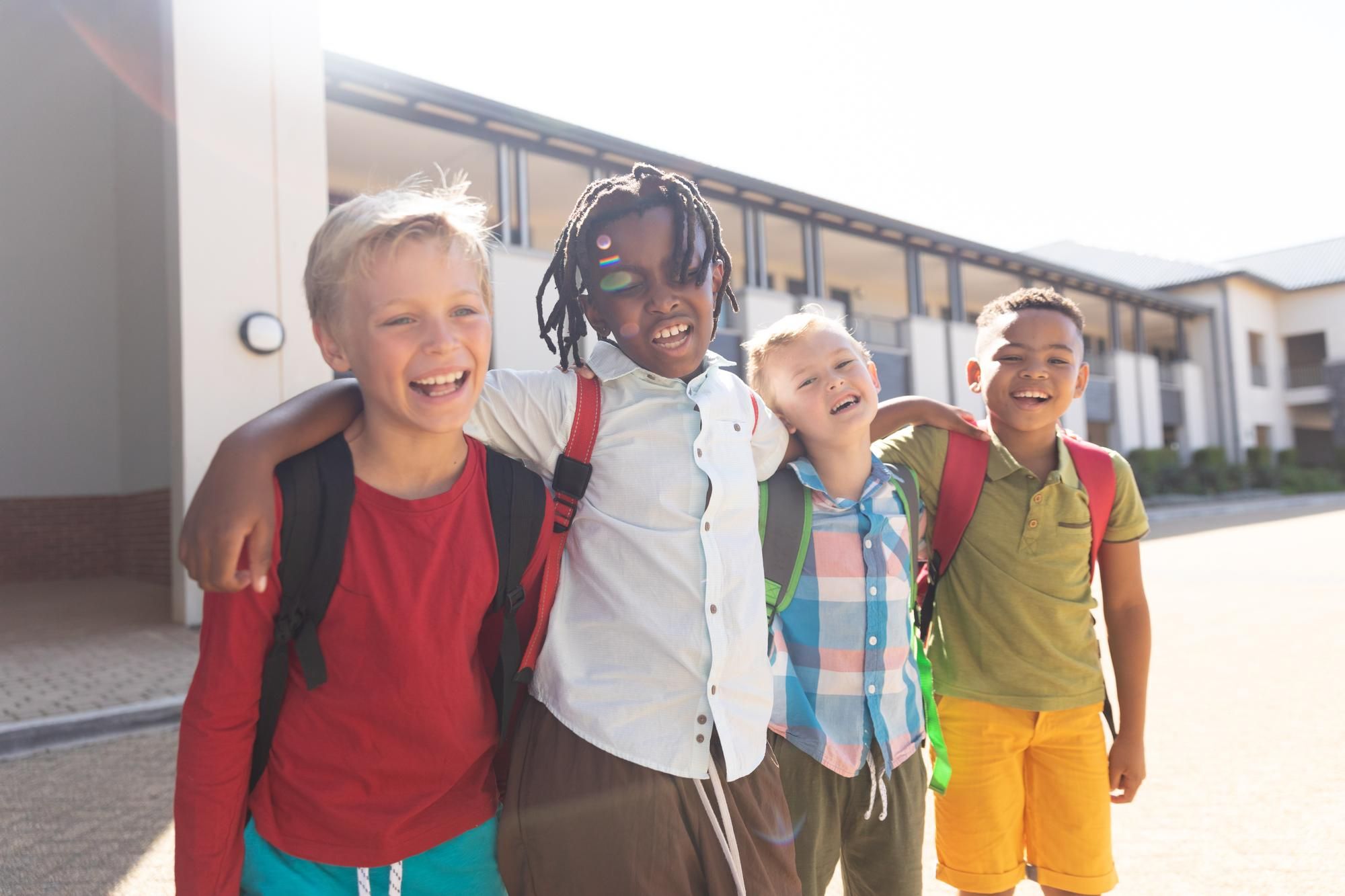 Four children with backpacks smile and embrace each other outdoors, conveying friendship and joy against a sunny school building backdrop.