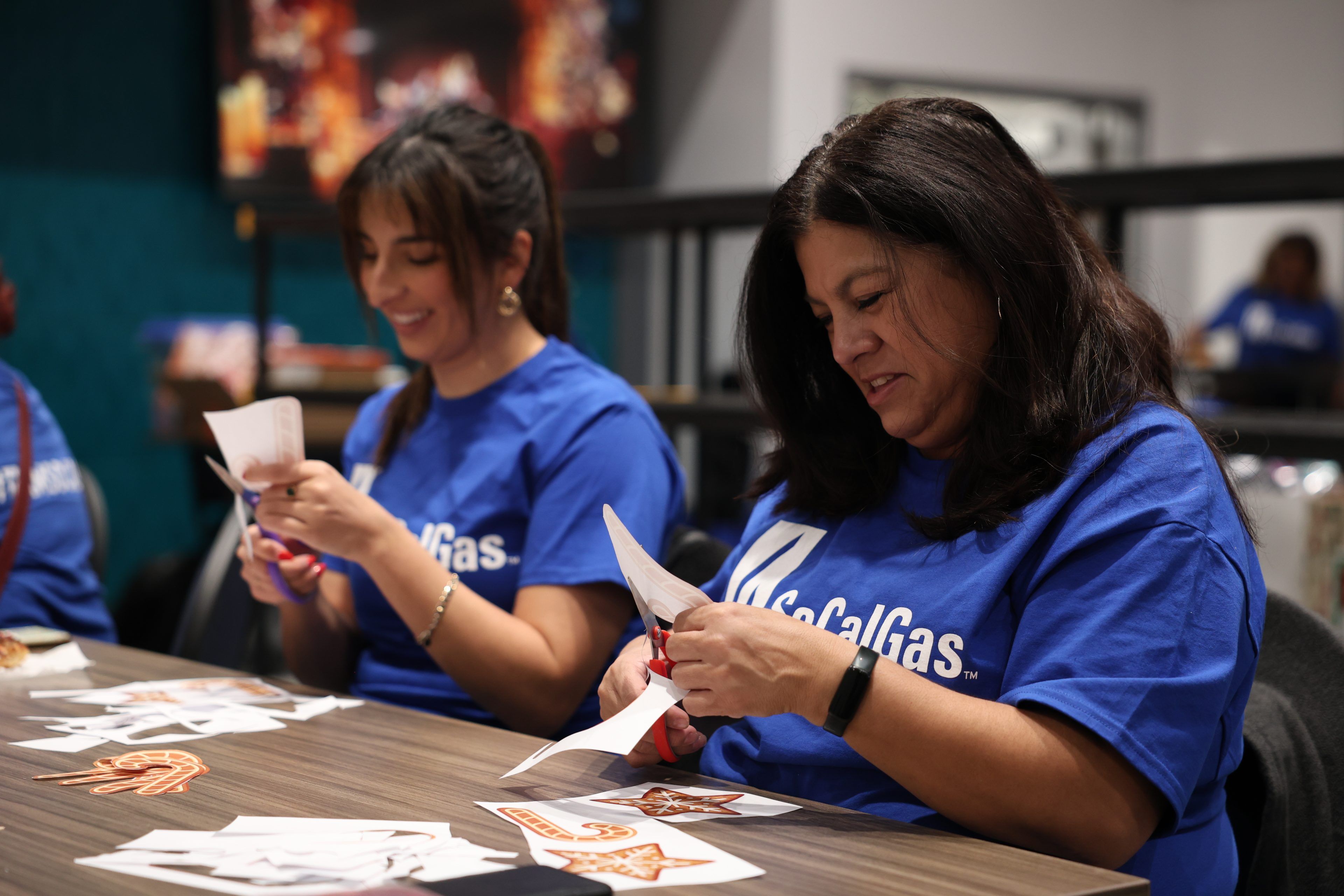 Two women in blue shirts sit at a table, smiling and concentrating as they cut paper. The setting appears relaxed and focused.