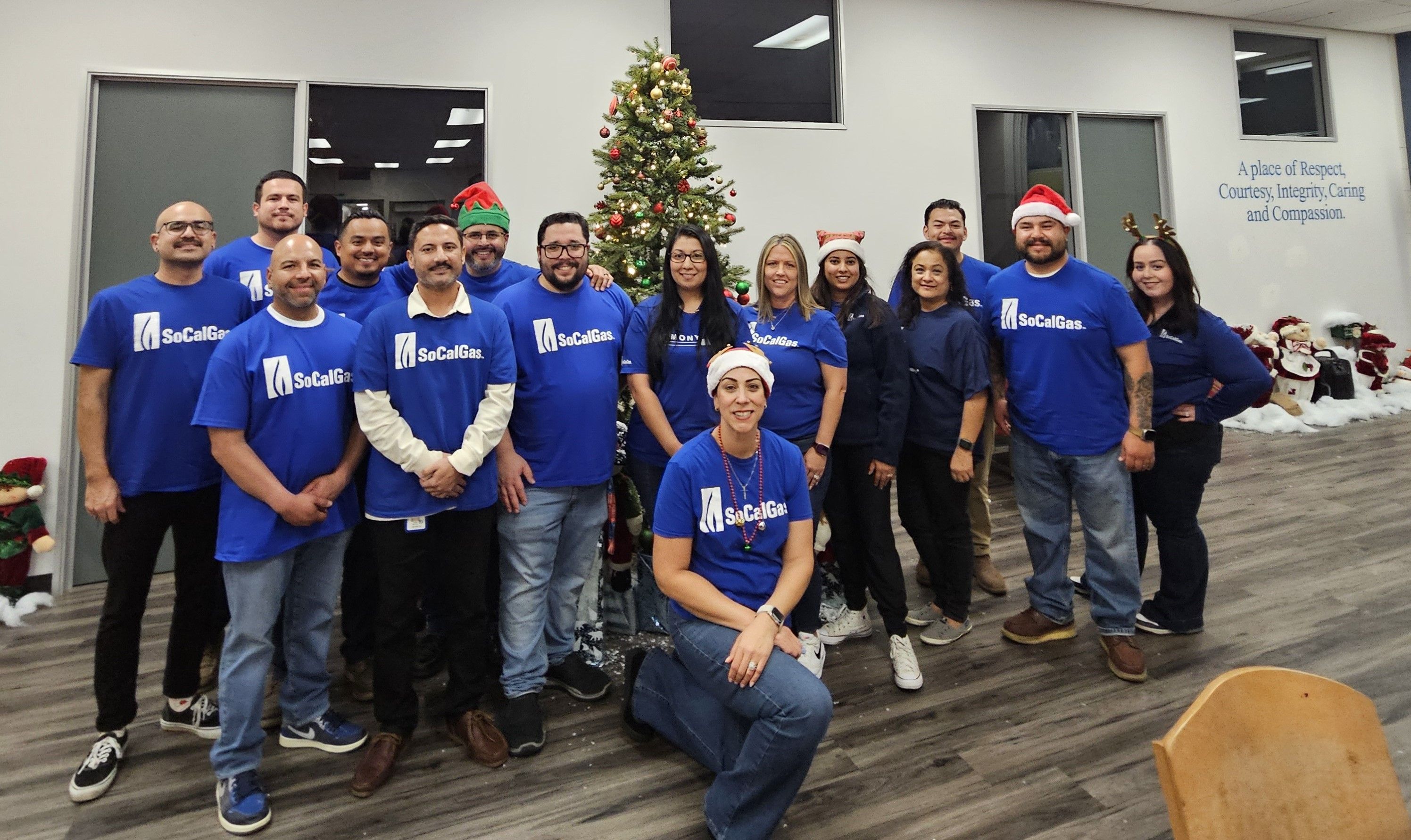 A group of people in blue shirts and festive hats smiles in front of a decorated Christmas tree in an office setting, conveying a joyful holiday spirit.