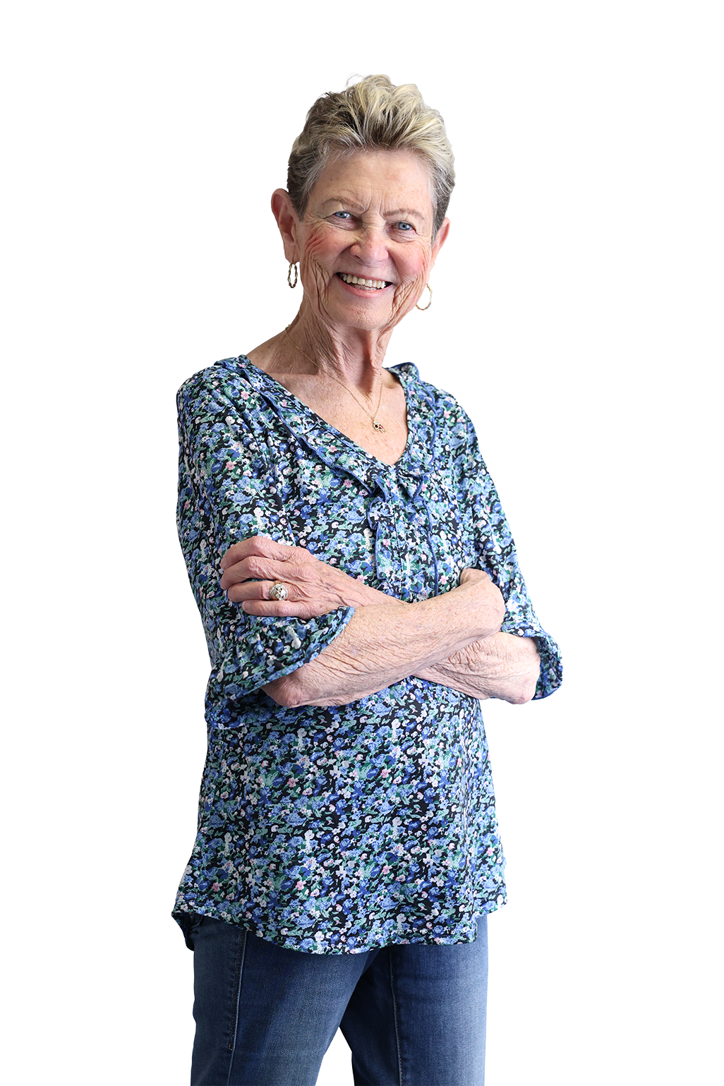 Smiling elderly woman with short hair, wearing a blue floral blouse and jeans, arms crossed, on a black background, conveying confidence and warmth.