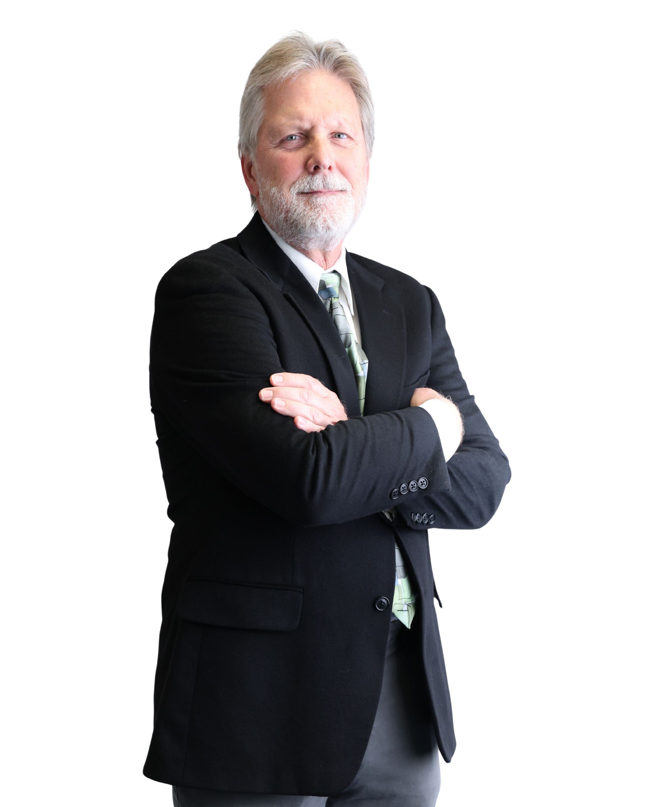 Elderly man in a black suit with crossed arms stands confidently against a black background. He has a light beard and wears a light-colored tie.