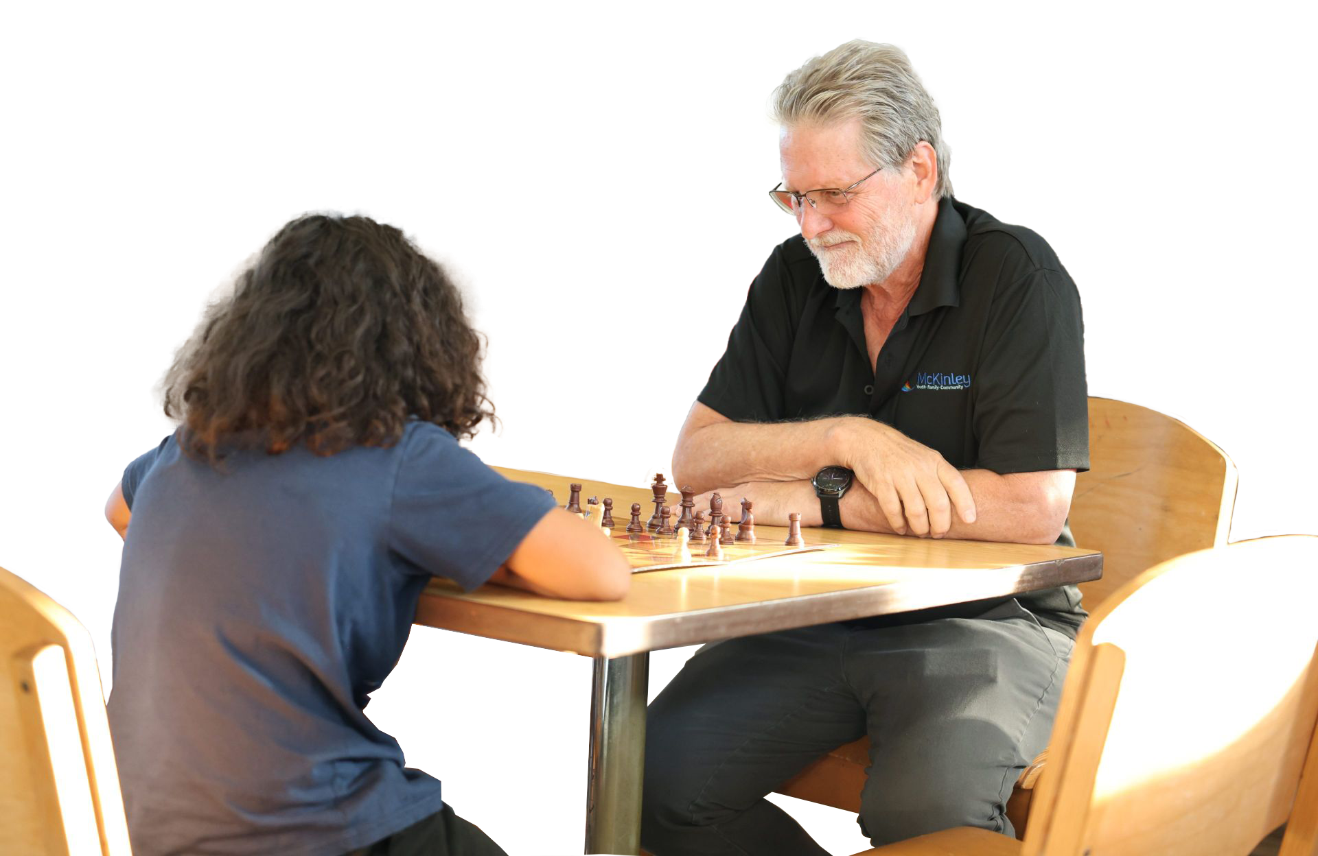 A gray-haired man and a young person intently play chess at a wooden table. The background is black, focusing entirely on the contemplative game scene.