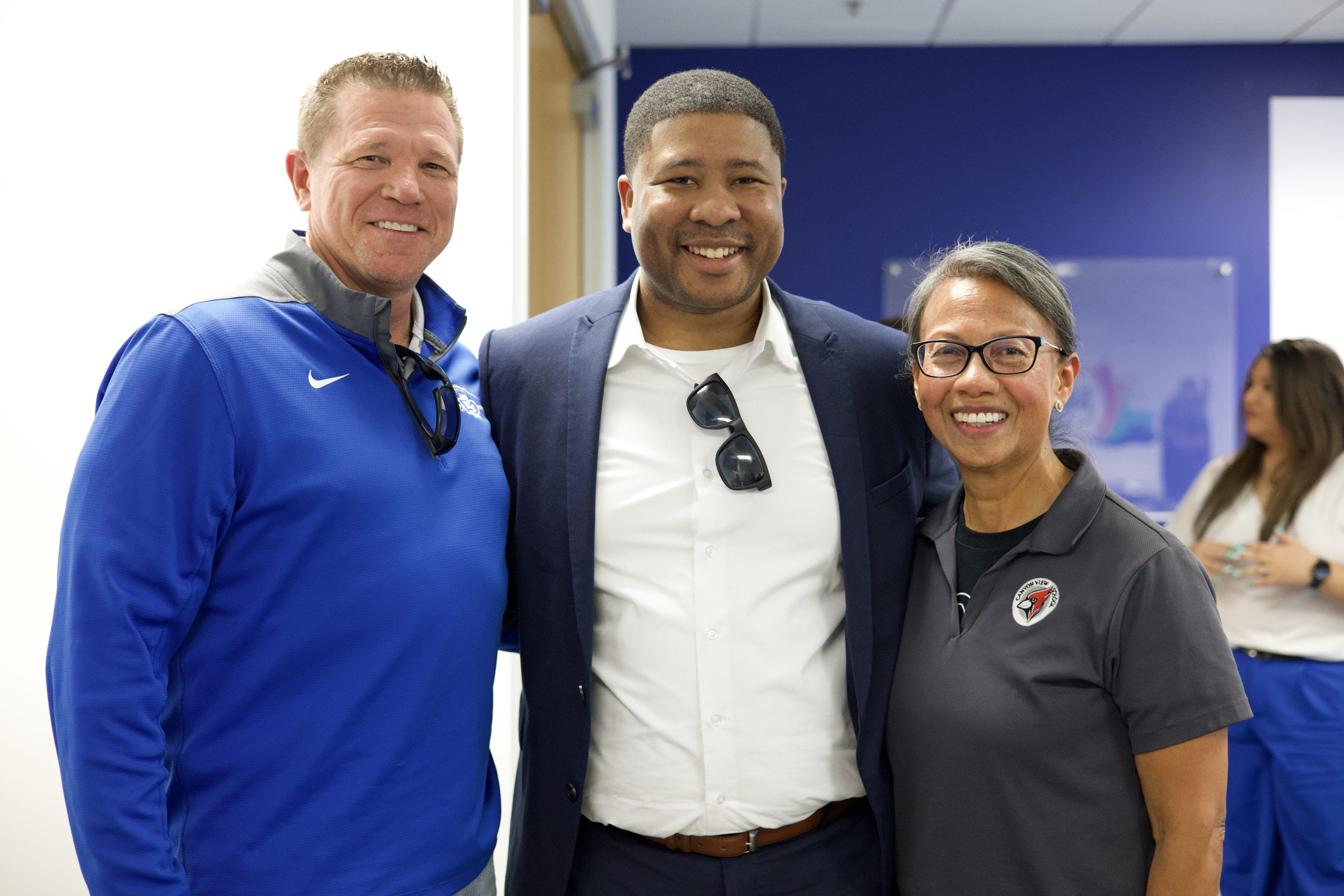 Three adults smiling, standing together indoors. The man on the left wears a blue jacket, the center man in a suit, and the woman on the right in a polo.