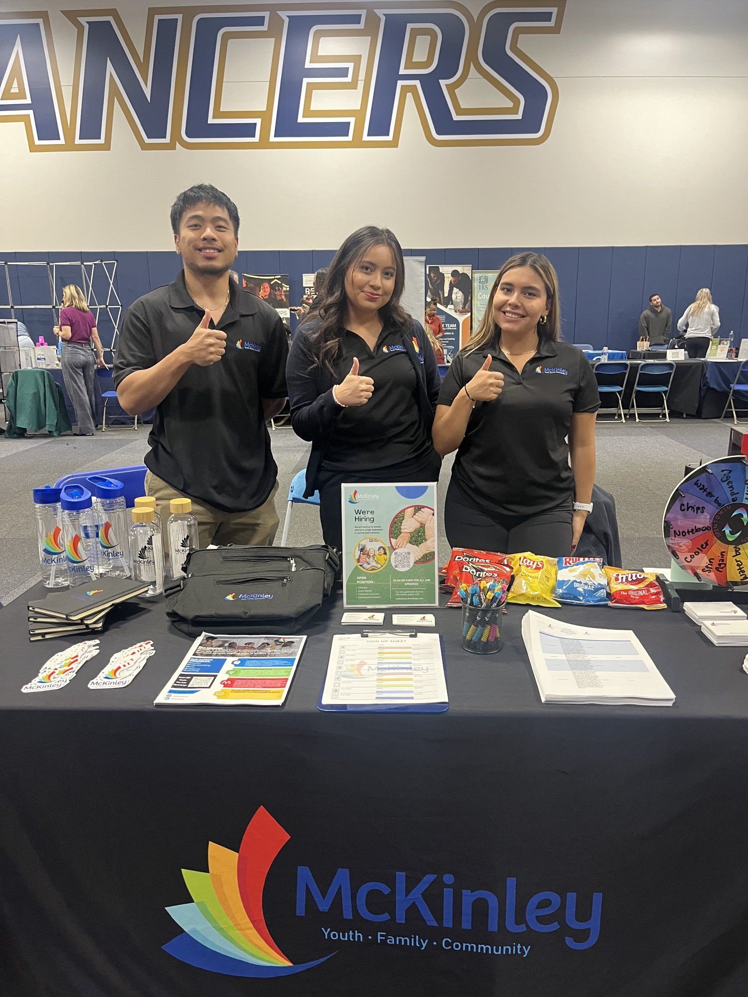 Three people stand behind a McKinley booth at a career fair, smiling and giving thumbs up. The table displays promotional items and snacks.