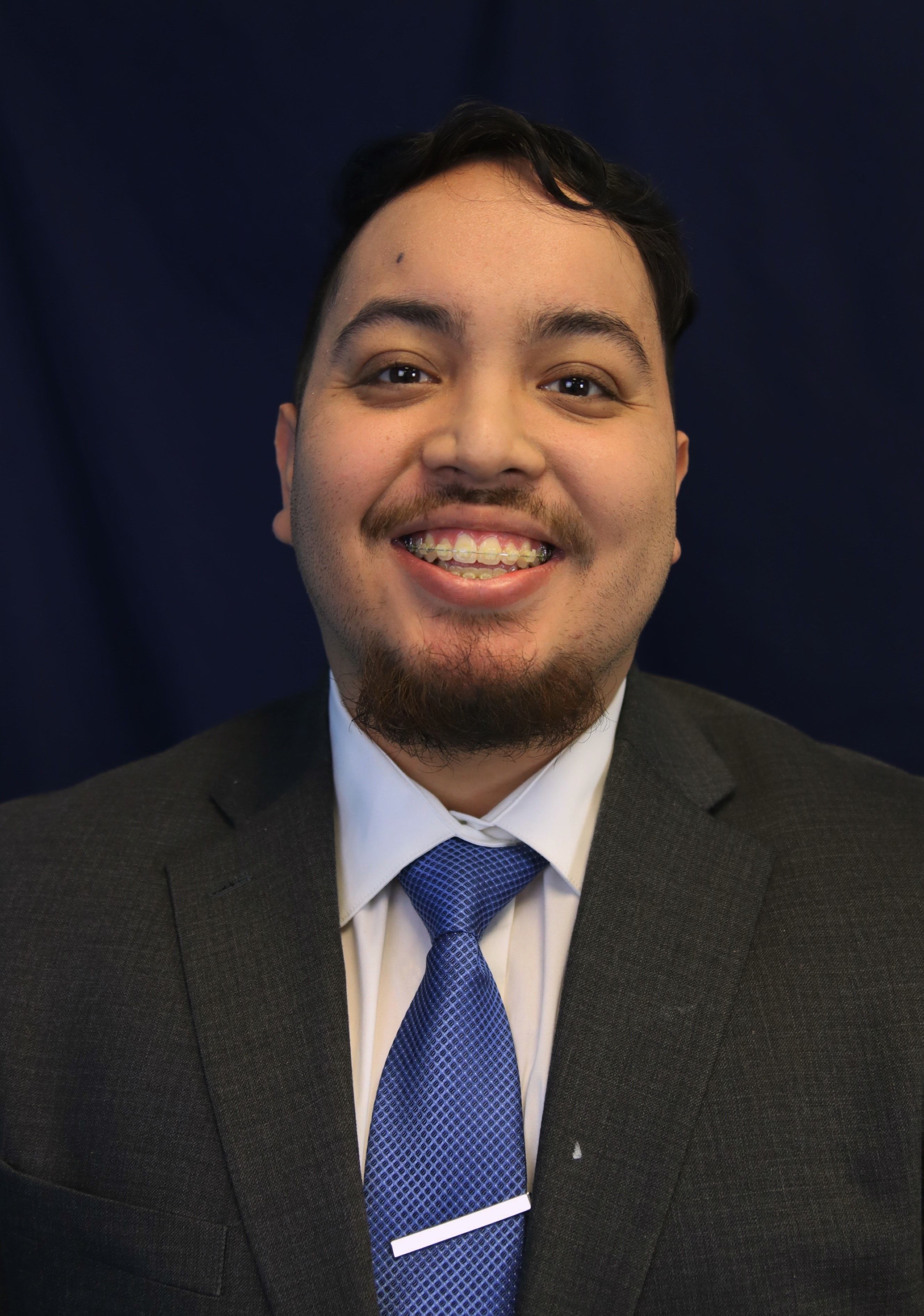 Smiling individual in a formal gray suit with a blue tie against a dark blue background, conveying a professional and approachable demeanor.