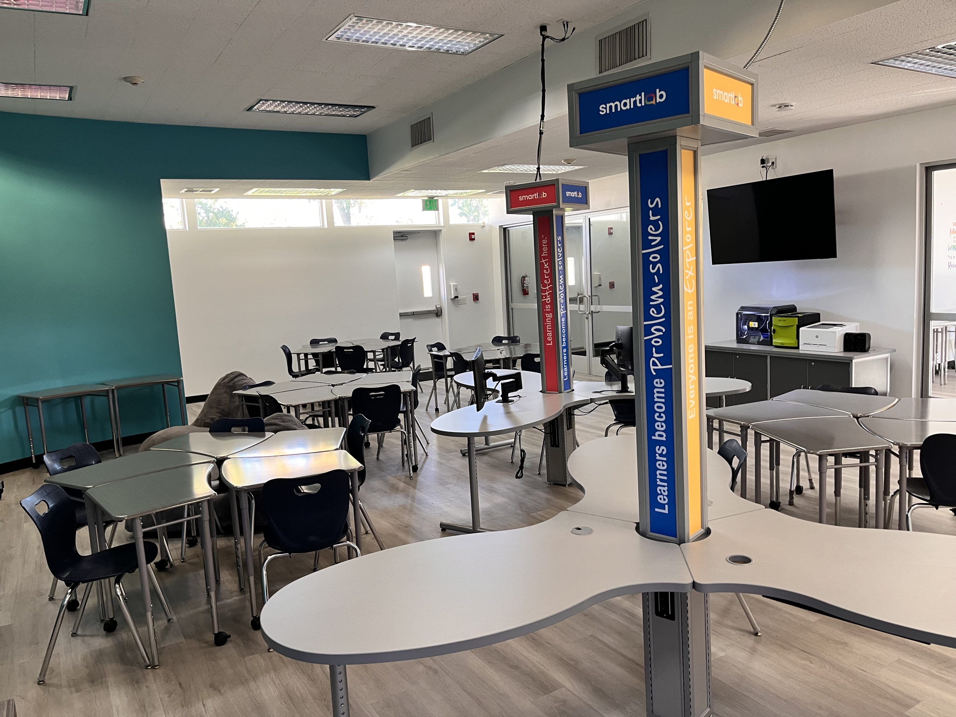 Modern classroom with colorful walls and flexible seating; tables and chairs scattered thoughtfully. A pillar displays "Learners become Problem-solvers."