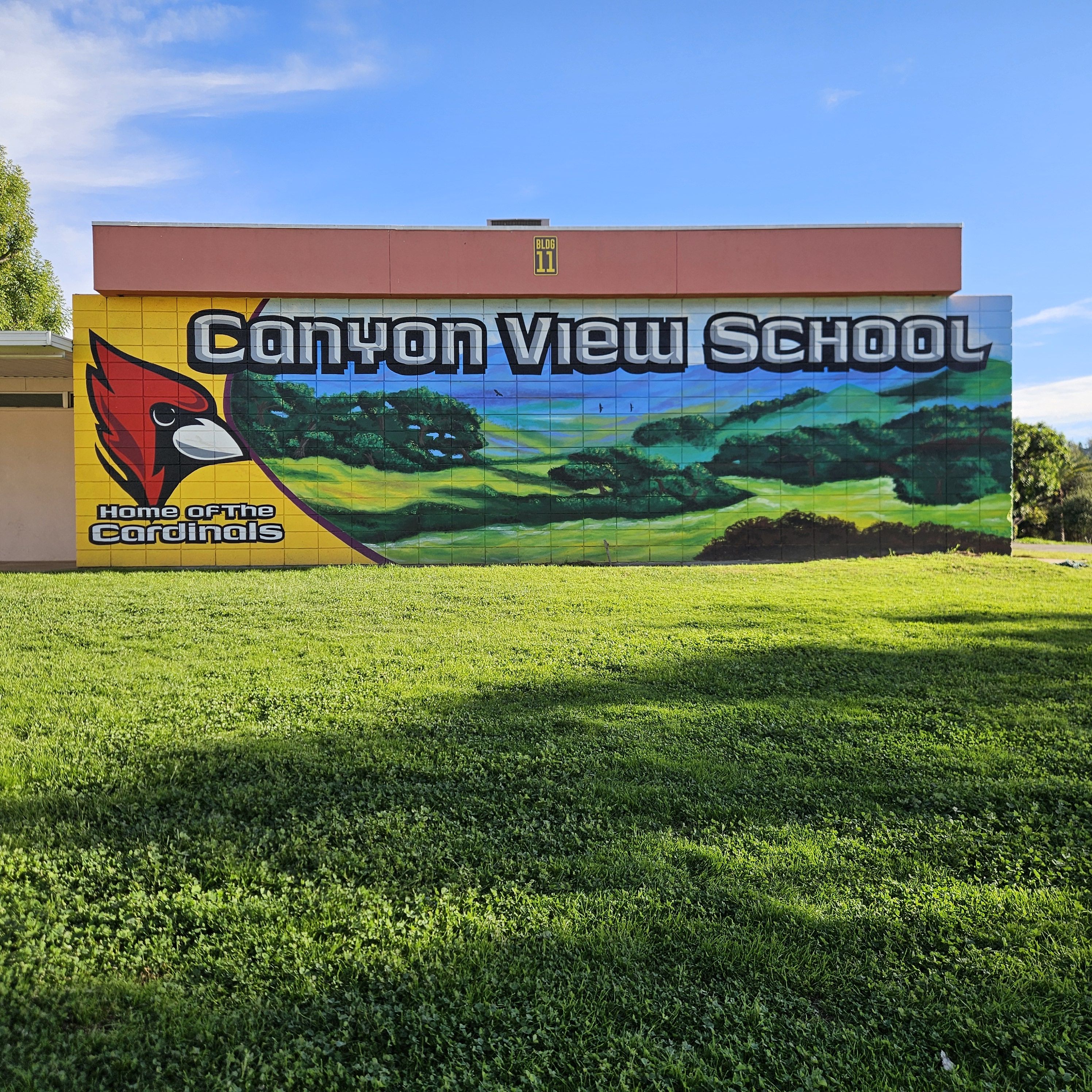 A mural on a building wall reads "Canyon View School" with a nature landscape scene, featuring a cardinal logo and the phrase "Home of the Cardinals." The foreground is a green lawn under a clear blue sky.