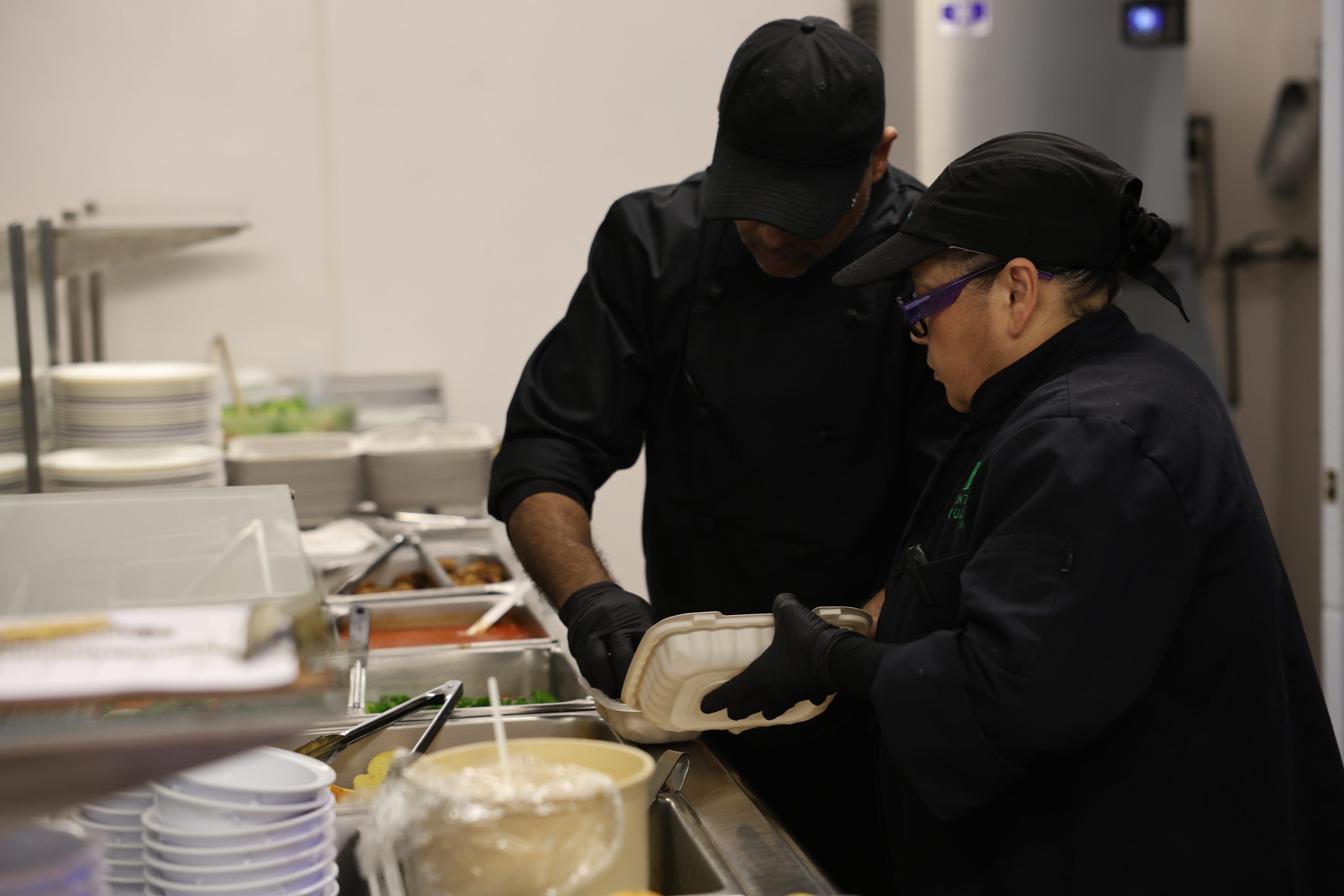 Two chefs in black uniforms and caps work in a kitchen, focused on preparing food in a takeout container. The scene conveys teamwork and concentration.