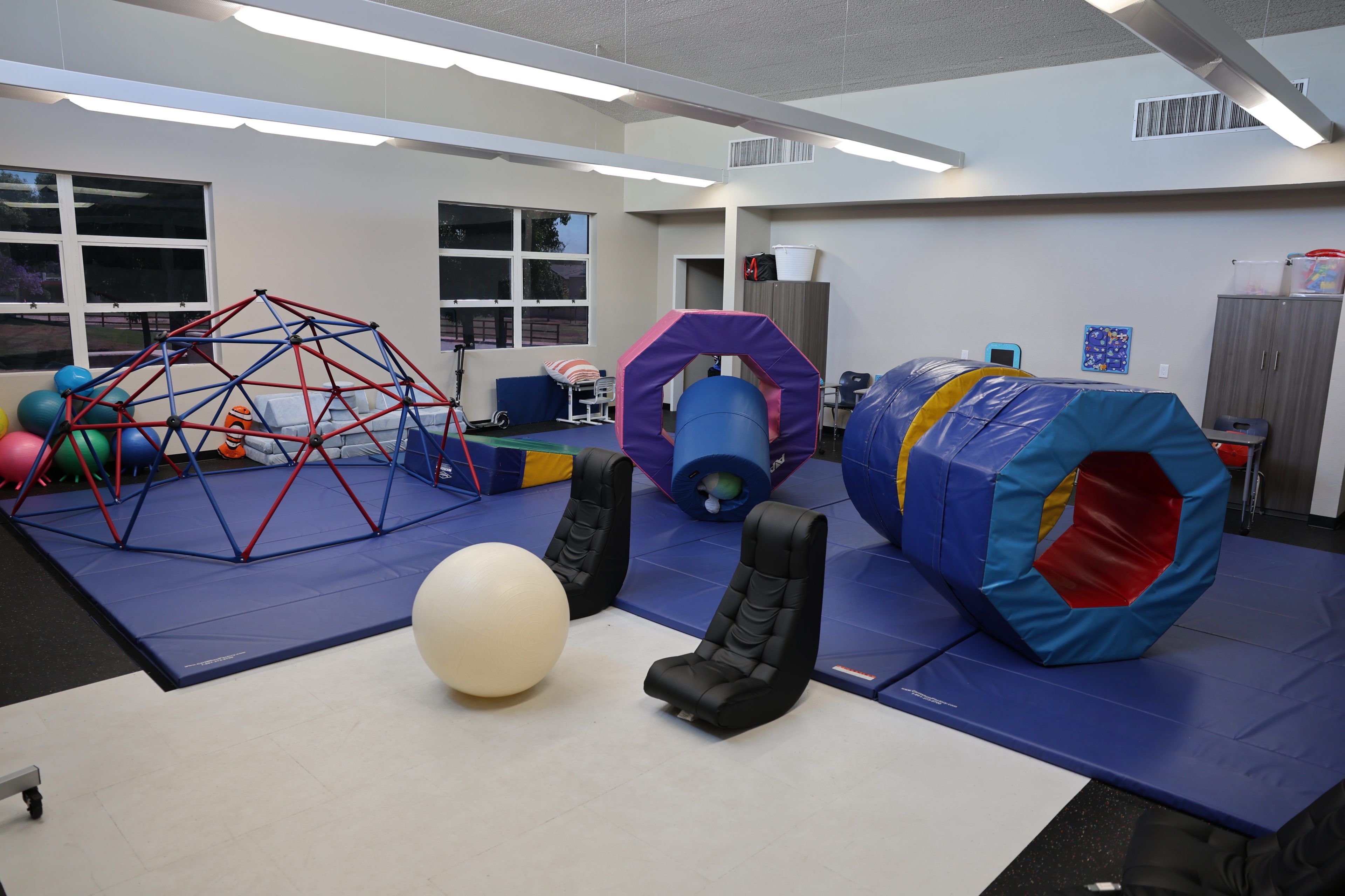 A children's play area with mats, geometric climbing structures, large cushions, exercise balls, and comfy chairs. The atmosphere is playful and inviting.