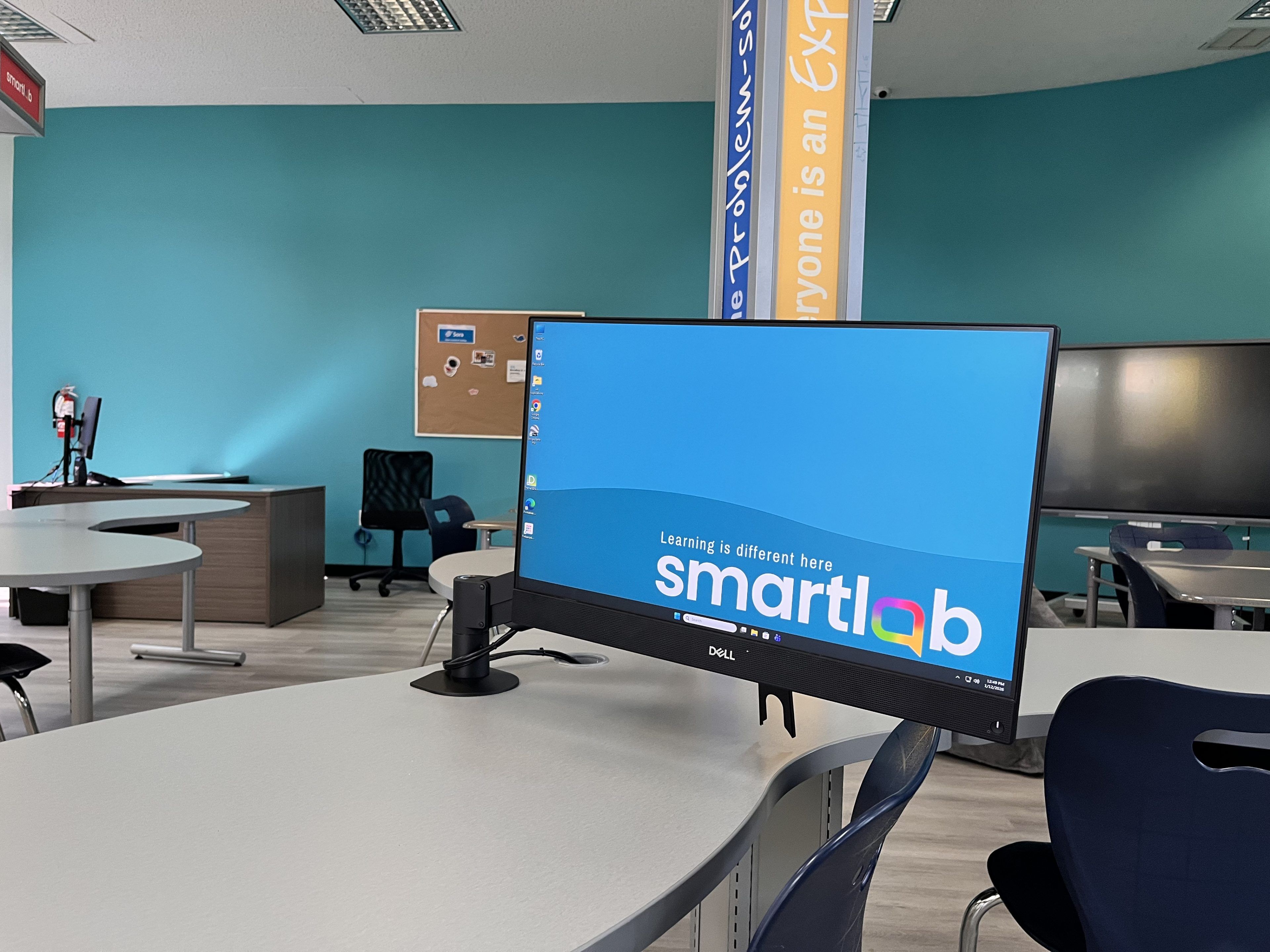 A modern classroom with a large monitor displaying "smartlab" on a desk. Blue walls, banners, and various seating create a tech-focused learning environment.