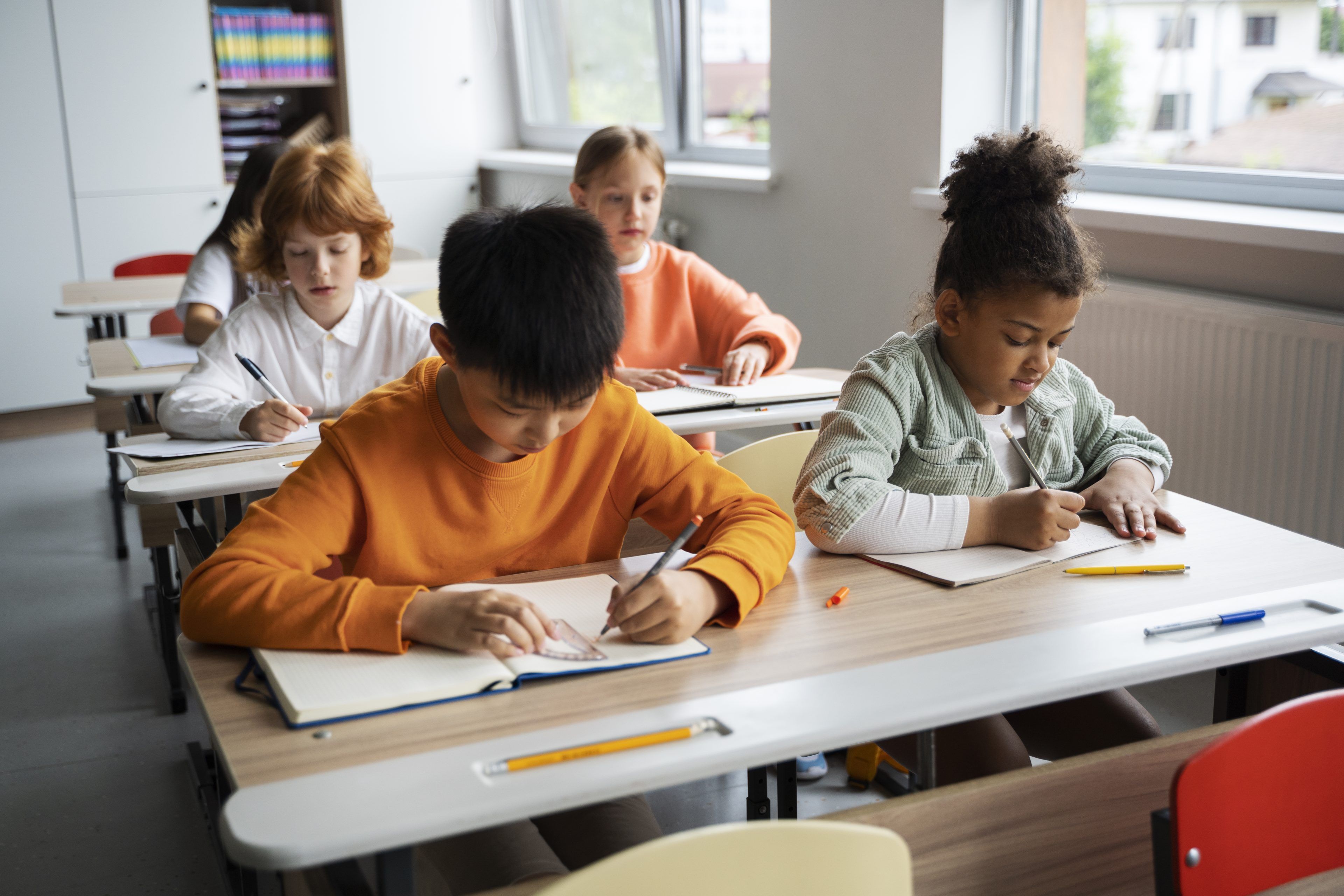 Four children sit at desks in a bright classroom, focused on writing in notebooks. The atmosphere is studious and calm, with pencils and books on desks.