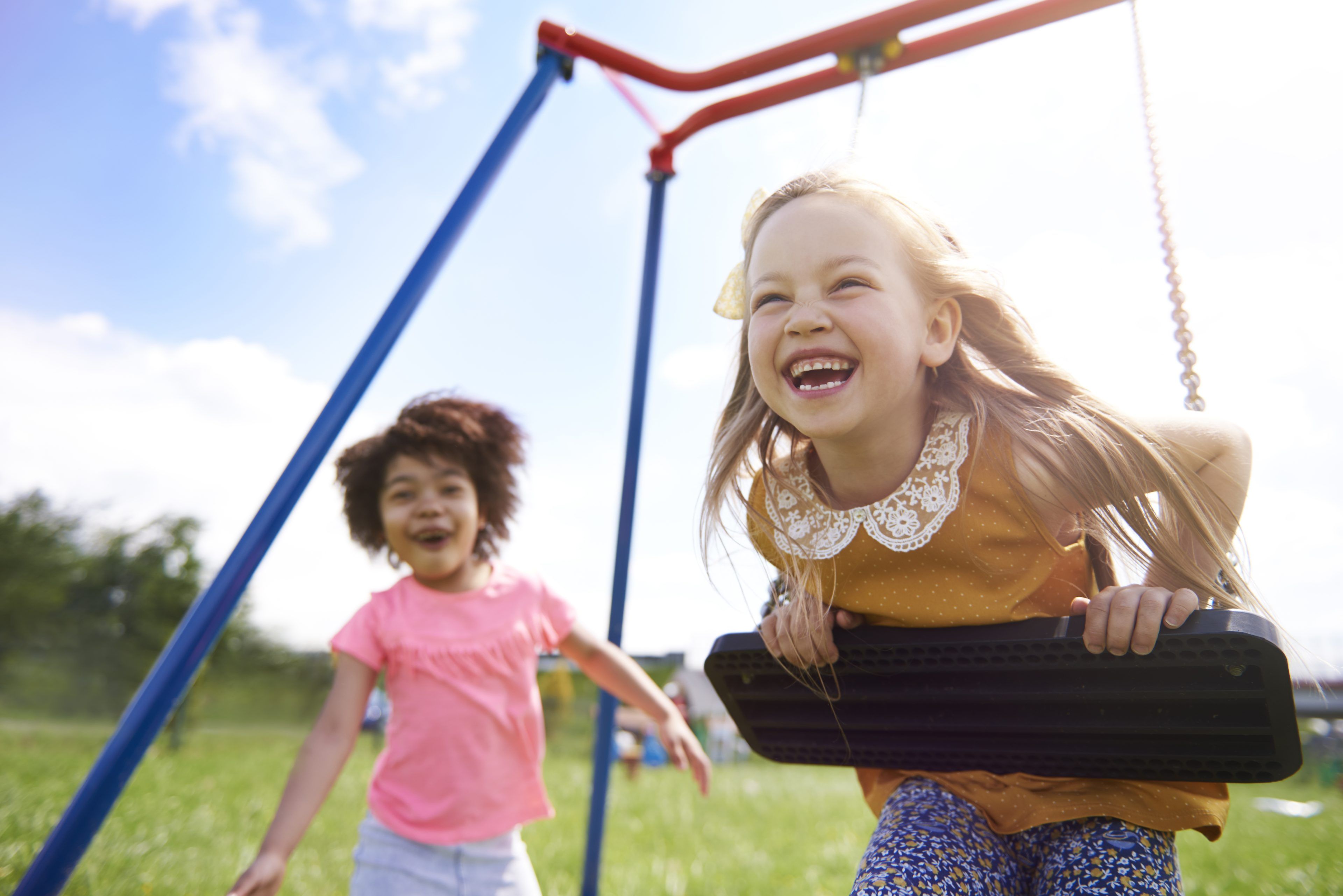 Two children play joyfully on swings in a sunny park. The girl in the foreground laughs while swinging high, radiating happiness. Bright blue sky above.