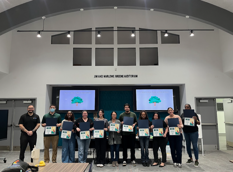 A group of twelve people stands in an auditorium, holding certificates and smiling. The atmosphere is celebratory, with "Jim and Marlowe Greene Auditorium" displayed above.