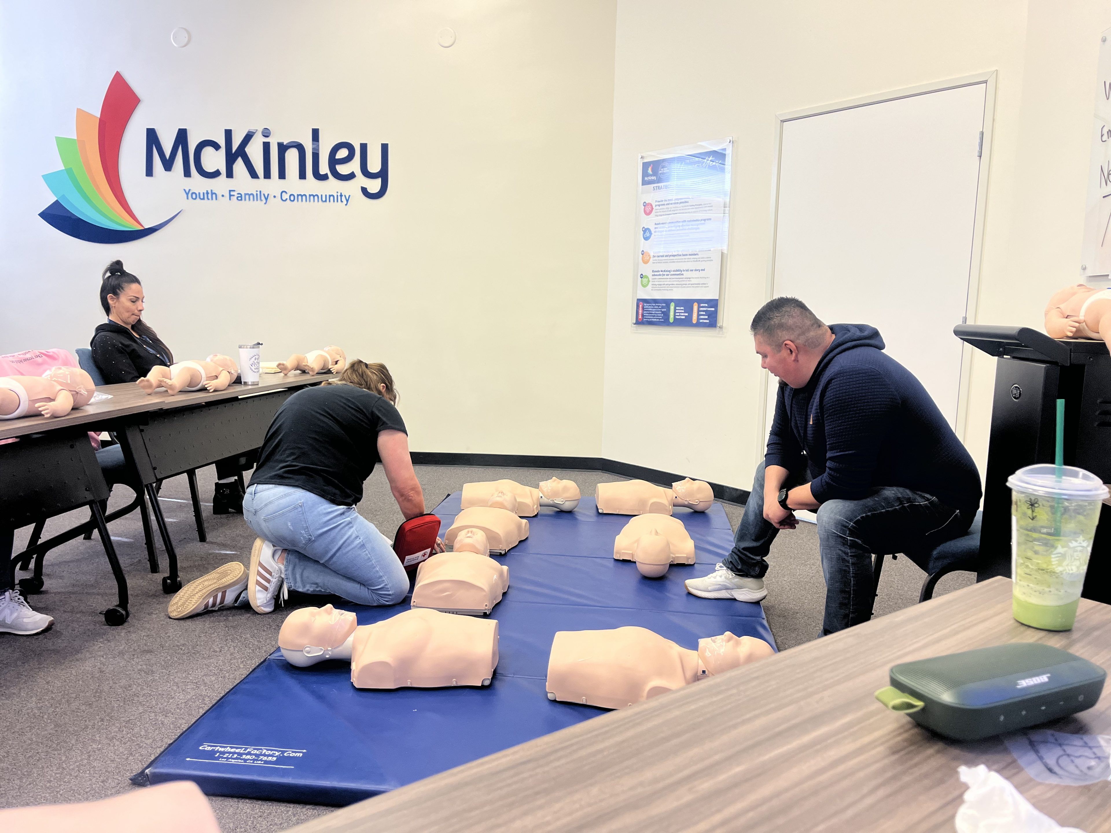 People practice CPR on mannequins in a classroom with "McKinley" on the wall. The scene conveys focus and learning. A poster and tables are visible.