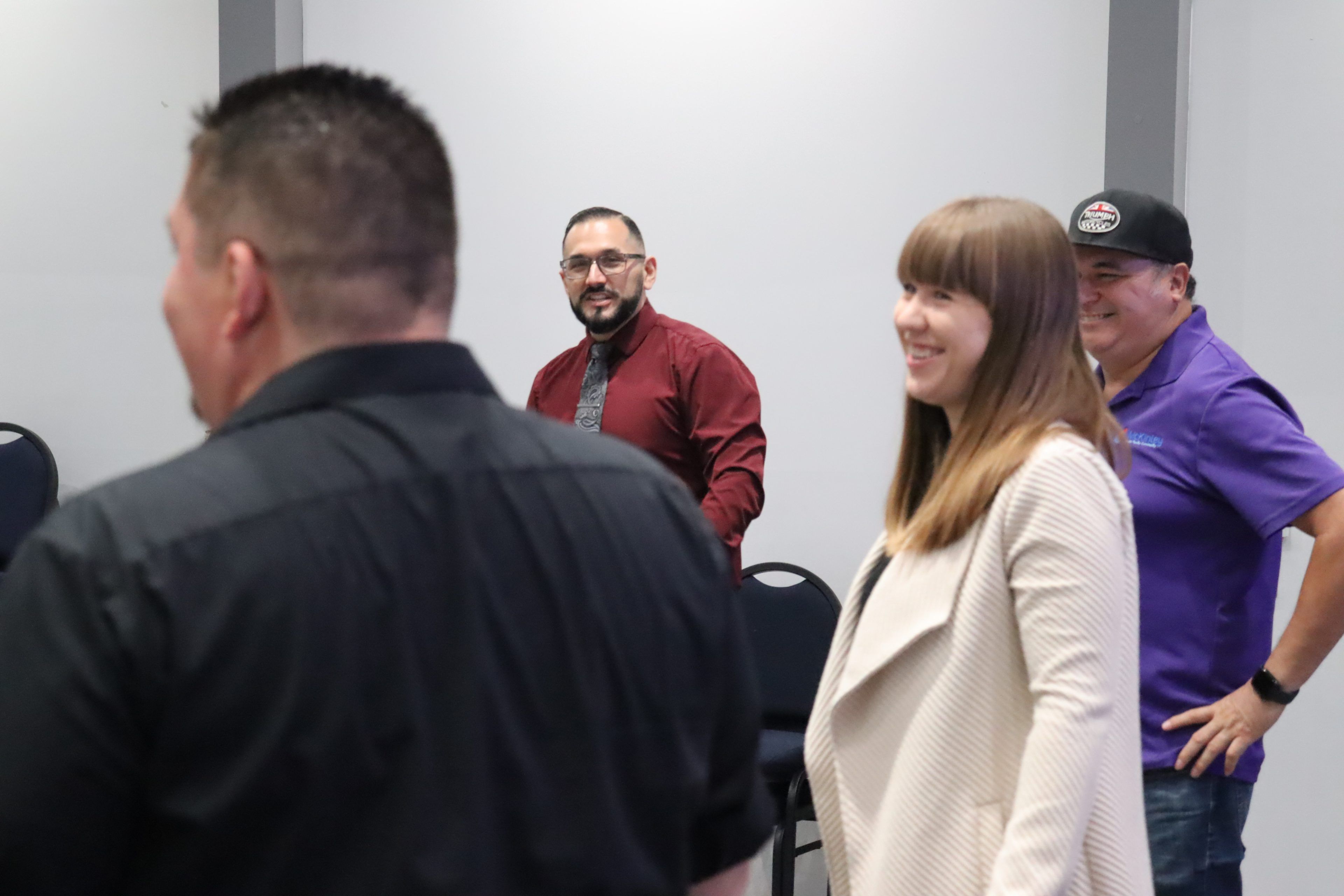 A group of people in a room are smiling and interacting. One person wears a maroon shirt, another is in a light jacket, and there's a visible sense of camaraderie.