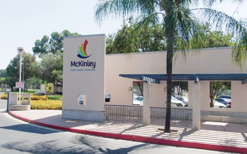 Small building with "McKinley" written on it, featuring a colorful logo. Surrounded by palm trees, clear sky, and a red-bordered sidewalk. Calm and inviting atmosphere.