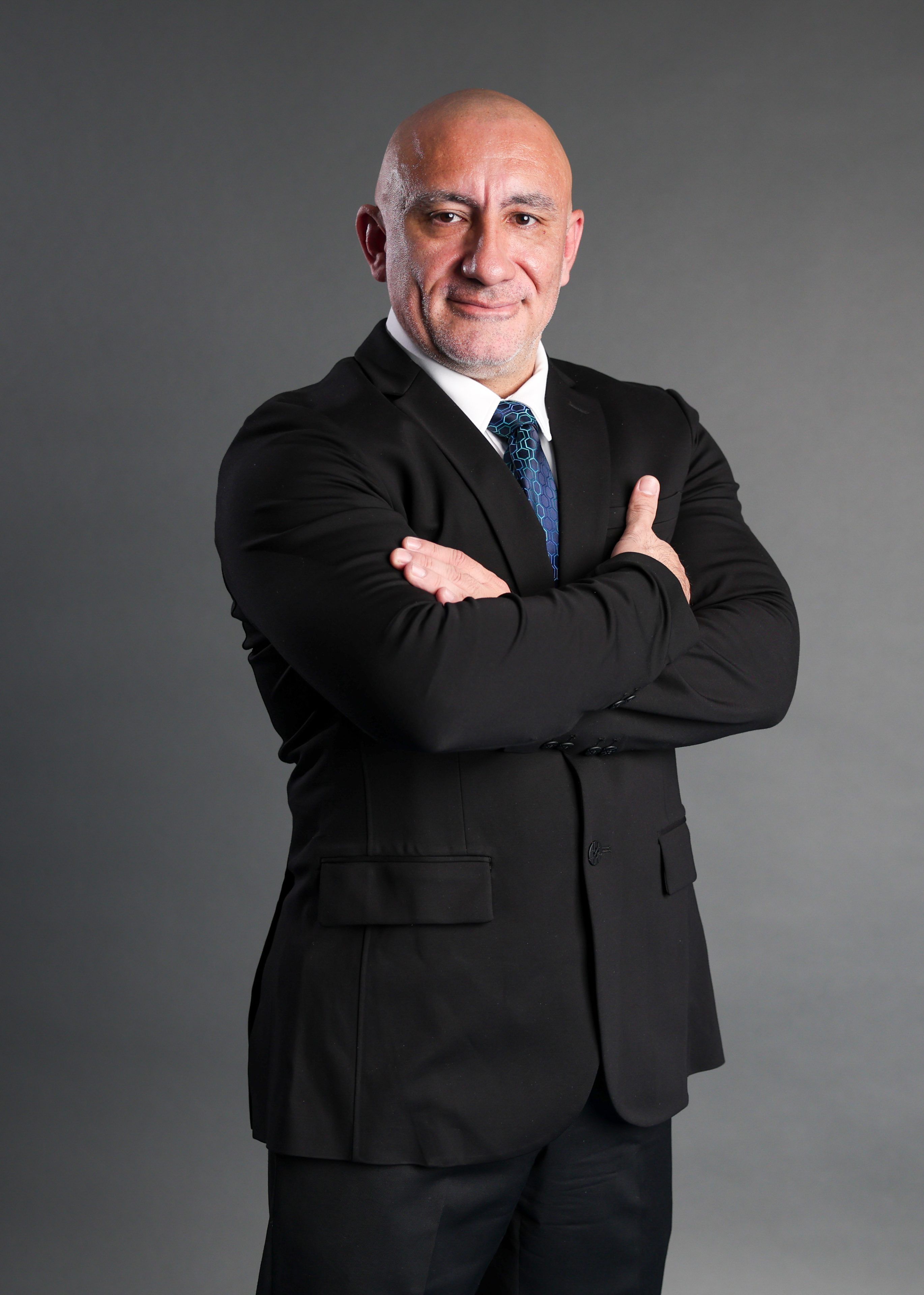 A bald man in a black suit and blue tie stands confidently with arms crossed against a gray background, conveying professionalism and authority.