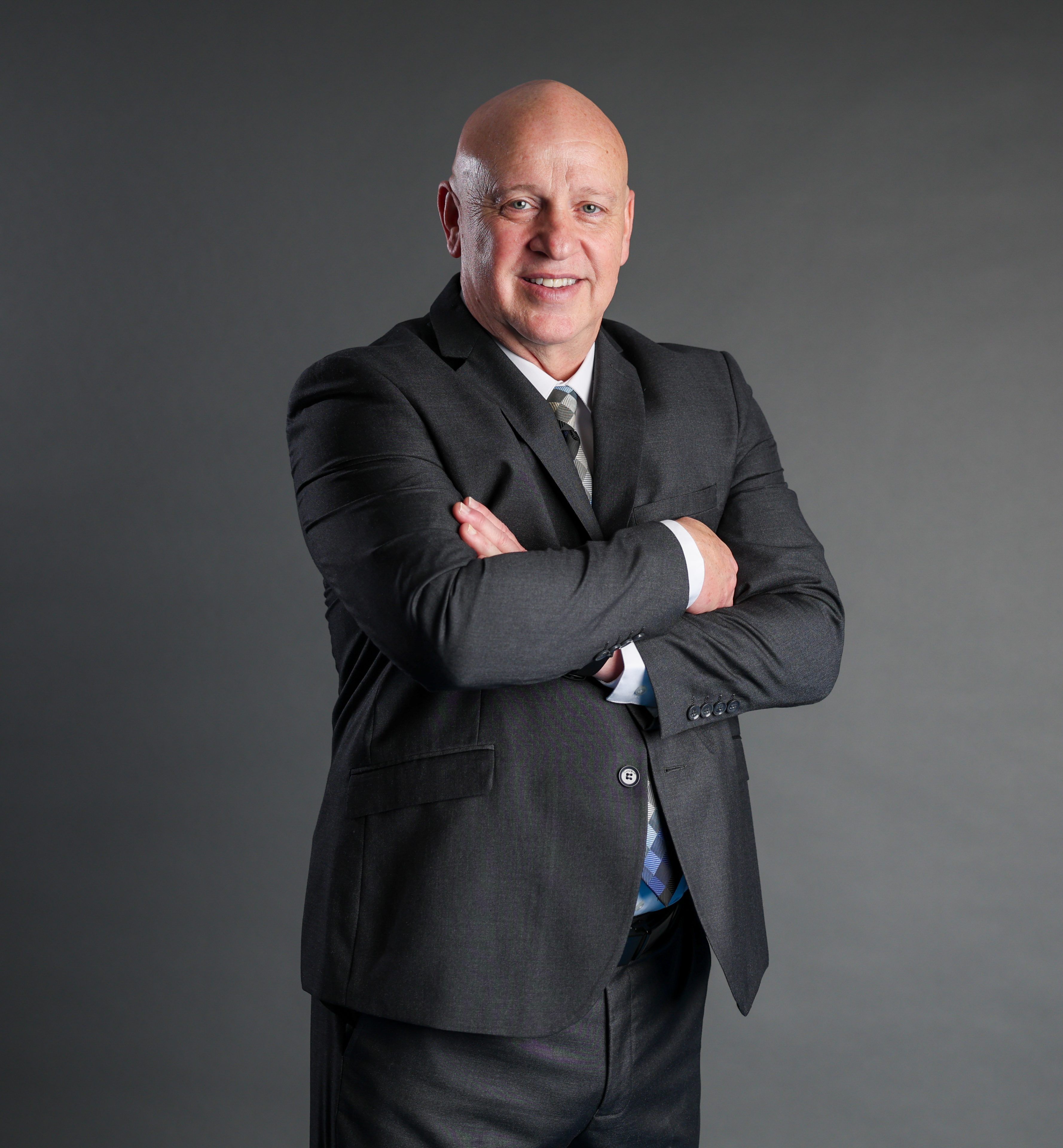 A confident bald man in a dark suit stands with arms crossed against a gray background, smiling warmly, conveying professionalism and approachability.