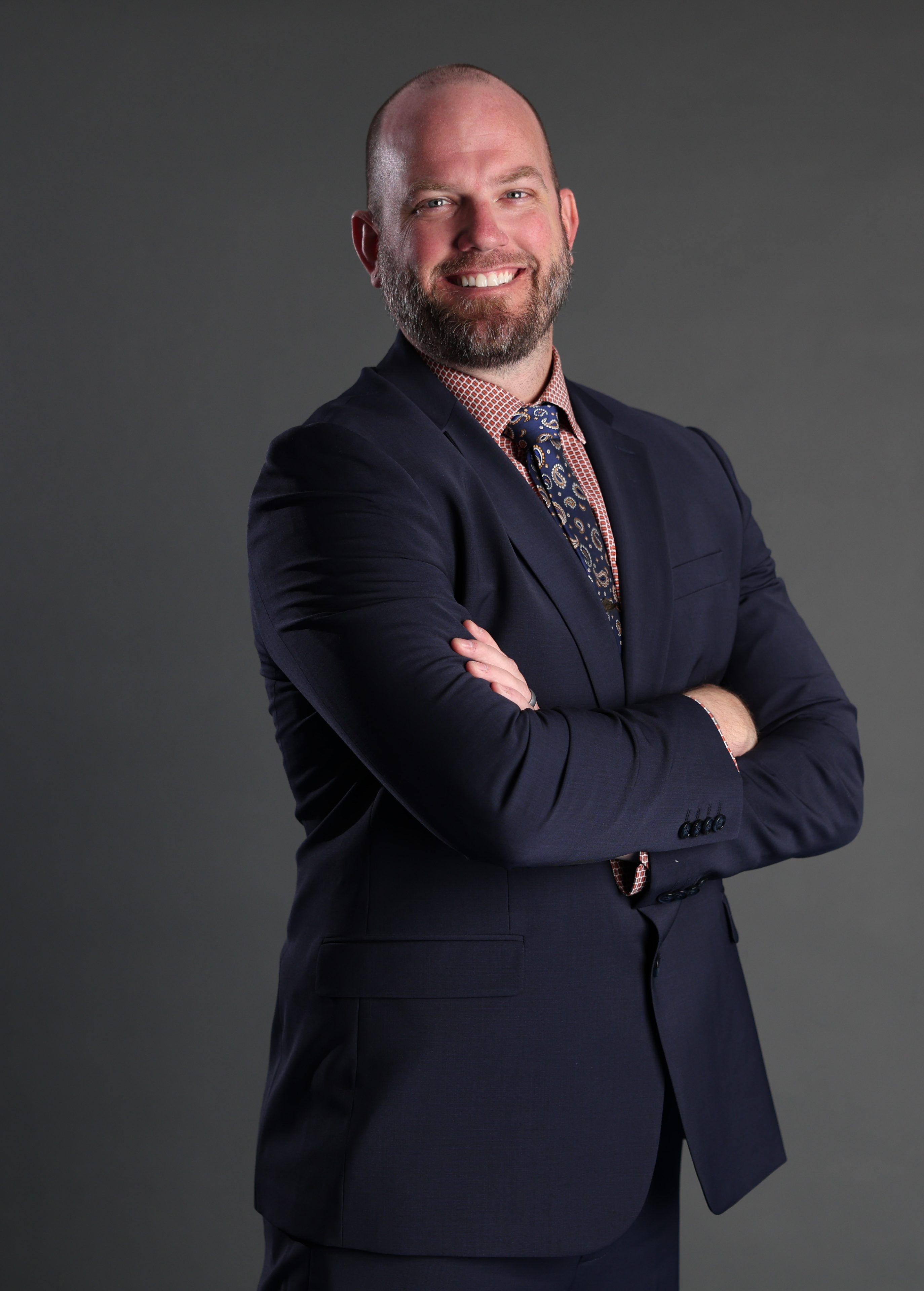 A smiling man with a beard stands confidently against a gray background, wearing a navy suit, patterned tie, and red-checkered shirt, arms crossed.