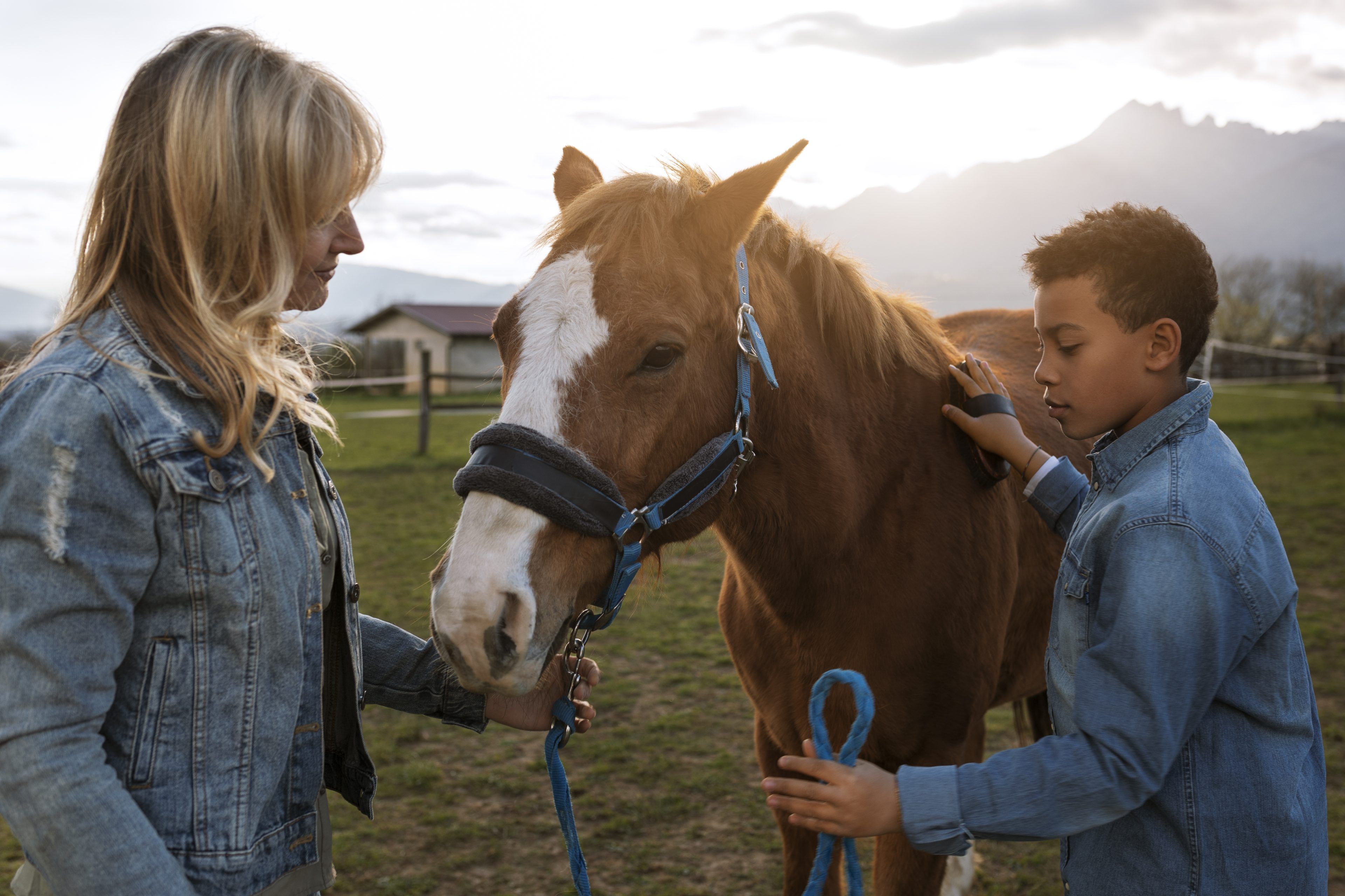 A woman and a young boy gently pet a brown horse with a white blaze on its face. They stand in a grassy field at sunset, both wearing denim jackets.