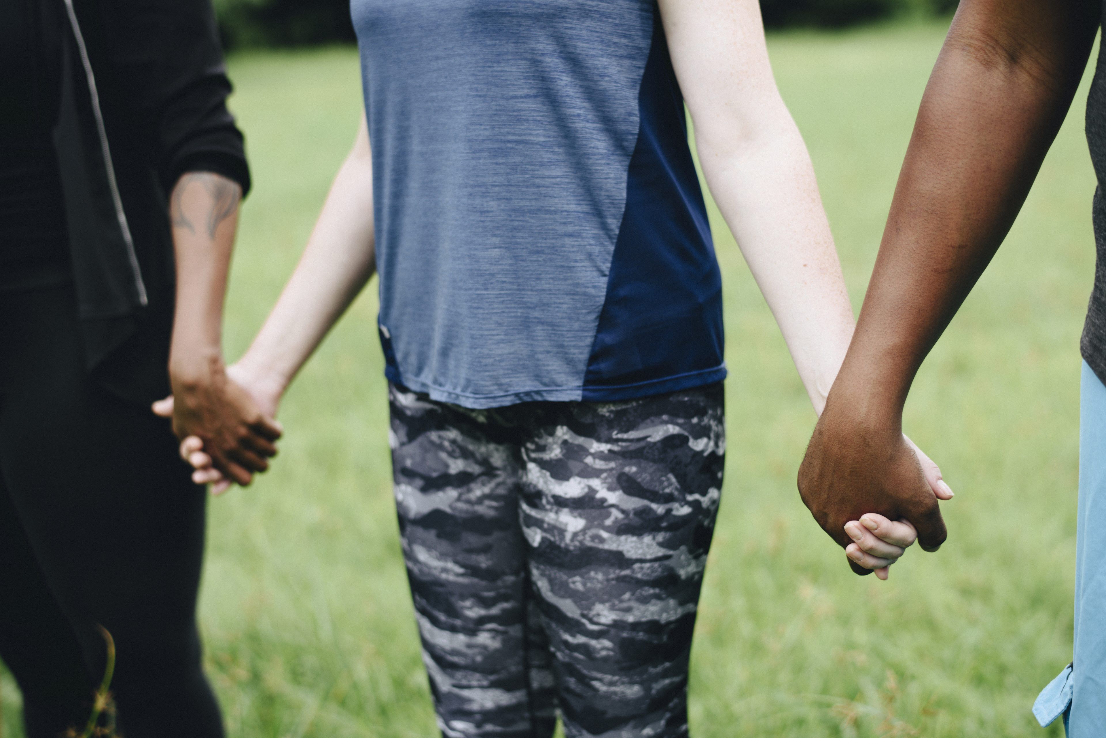 Three people holding hands outdoors, focusing on their joined hands and torsos. They stand on grass, suggesting unity and diversity. Casual attire, peaceful setting.