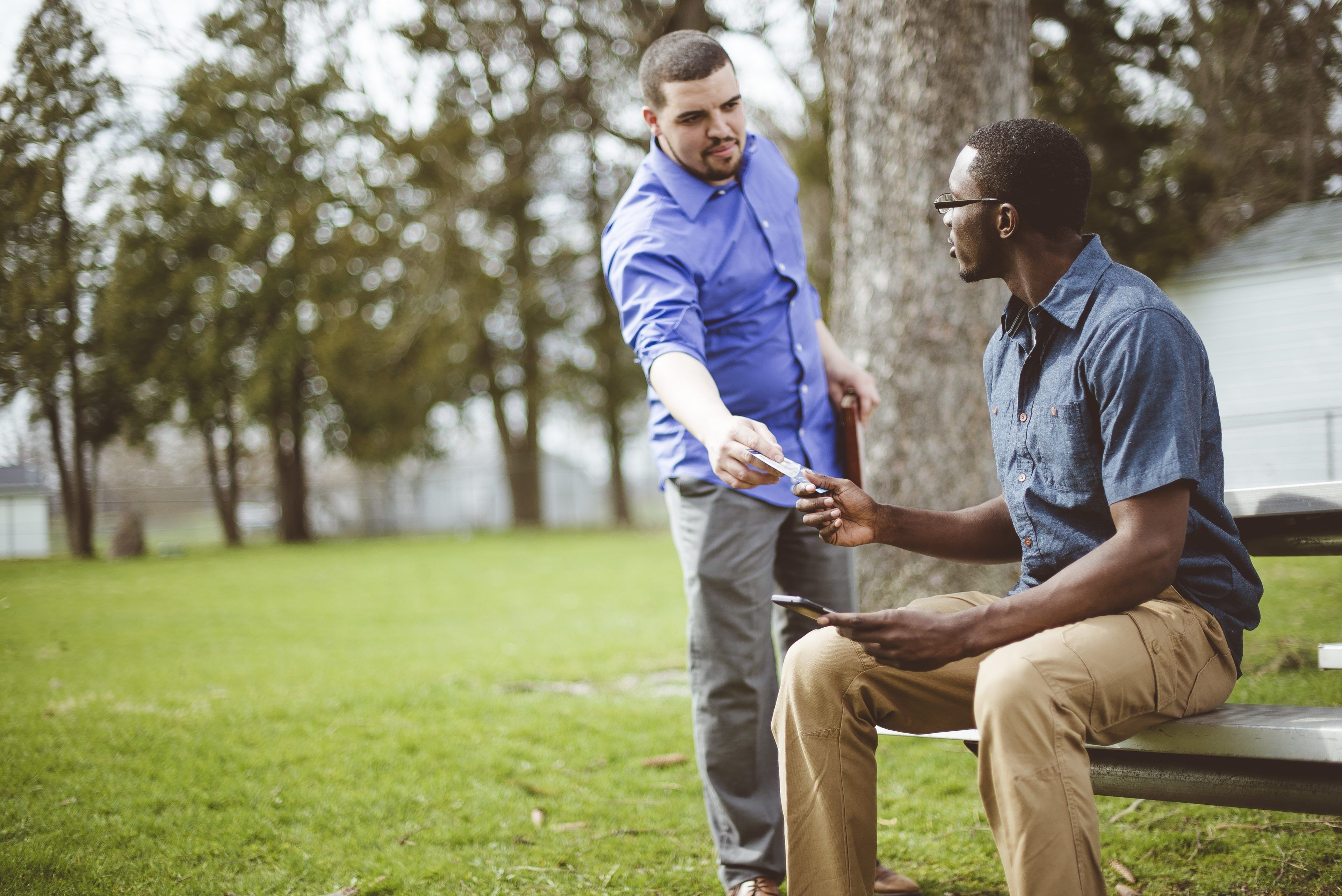 Two men in a park: one sitting on a bench accepting a smartphone from the other, who is standing. The scene is friendly and relaxed with trees in the background.