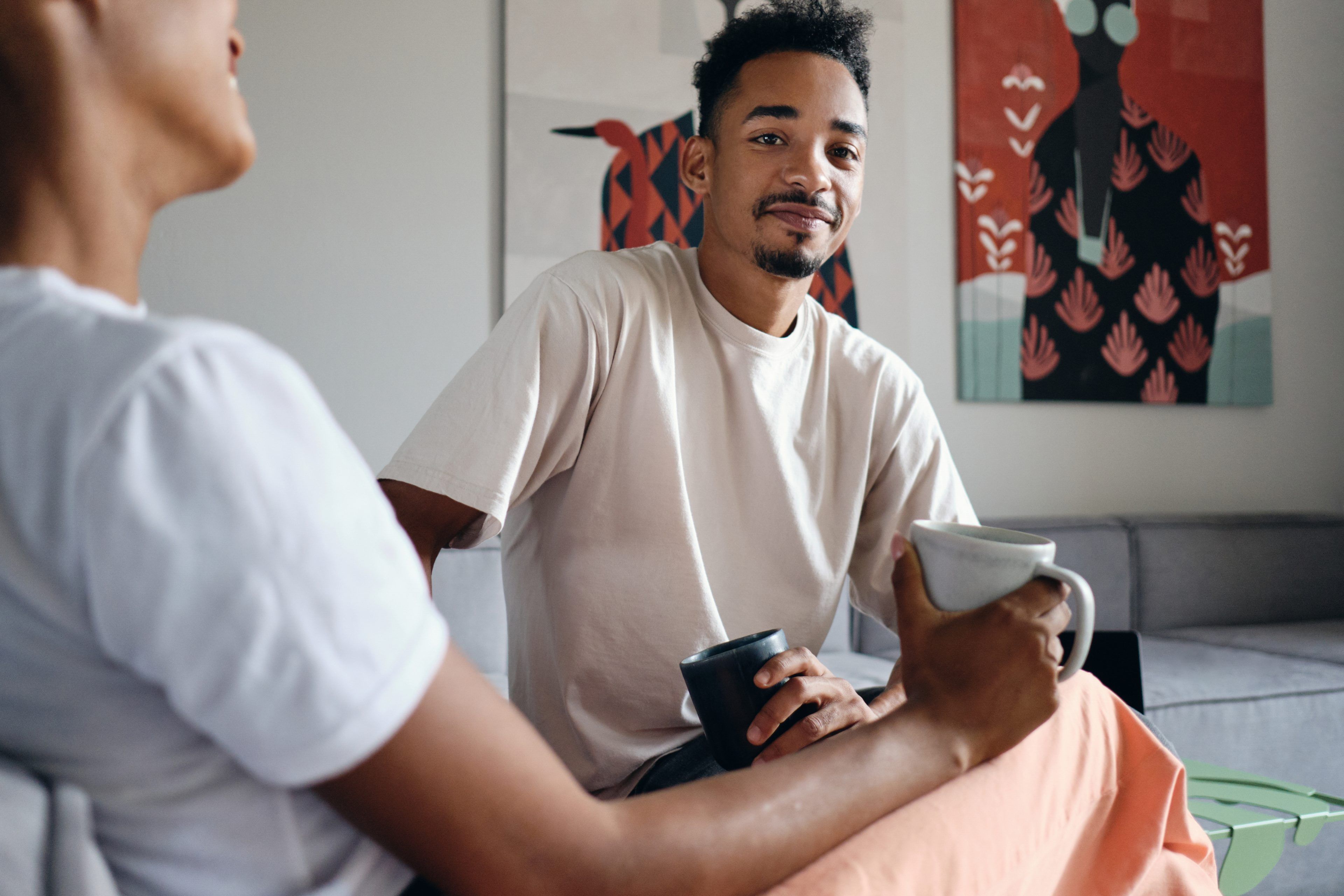 A man in a light shirt sits smiling, holding a mug, facing another person. Art on the walls in a cozy room suggests a relaxed, friendly atmosphere.